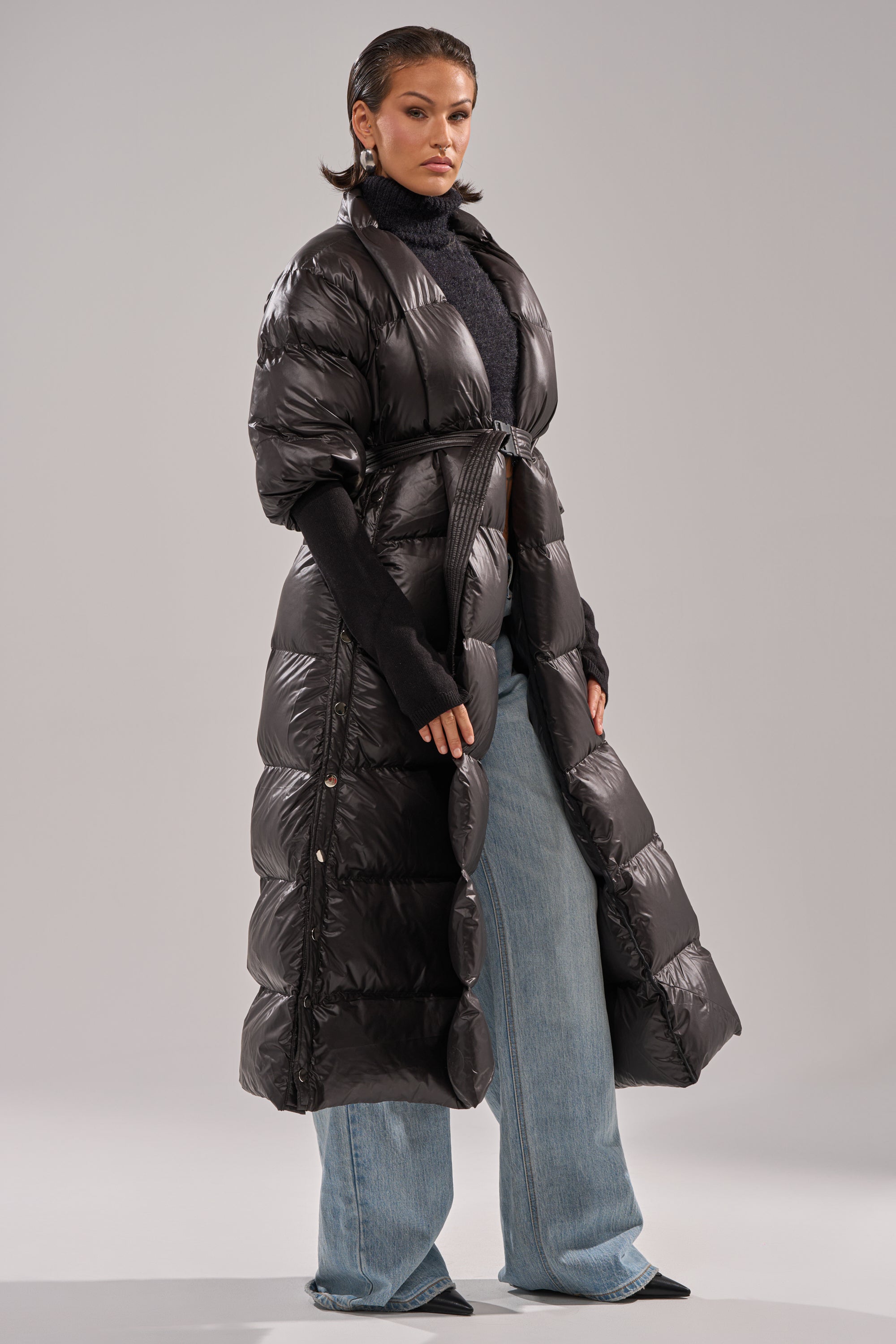 A woman stands confidently in the POPPIN OFF TEARAWAY PUFFER, a long, shiny black belted coat, layered over a dark turtleneck, flared light blue jeans, and black pointed-toe shoes against a plain gray background.