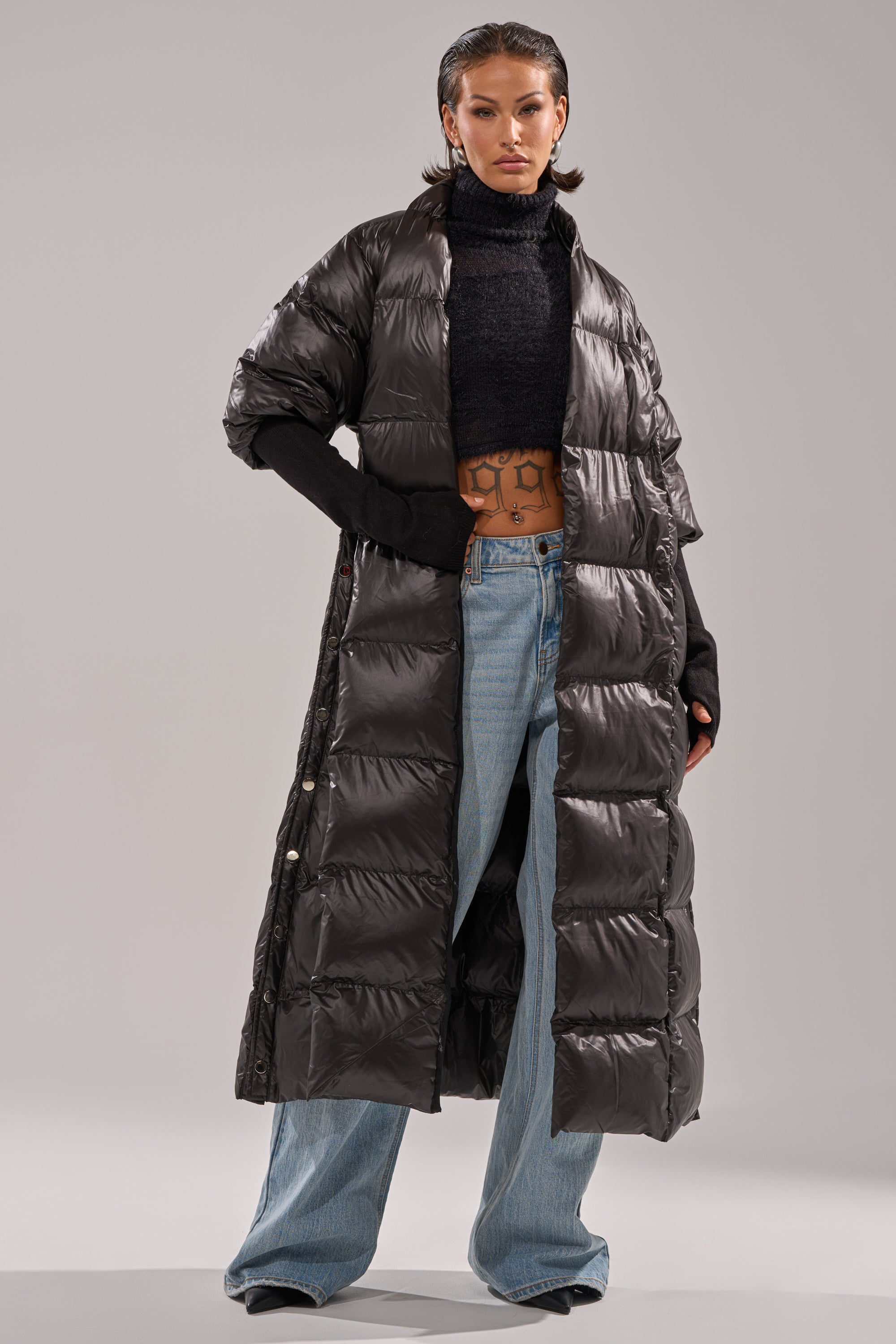A person wears the POPPIN OFF TEARAWAY PUFFER—a long, shiny black puffer coat—over a black cropped turtleneck, light blue jeans, and black gloves, standing against a plain gray background.