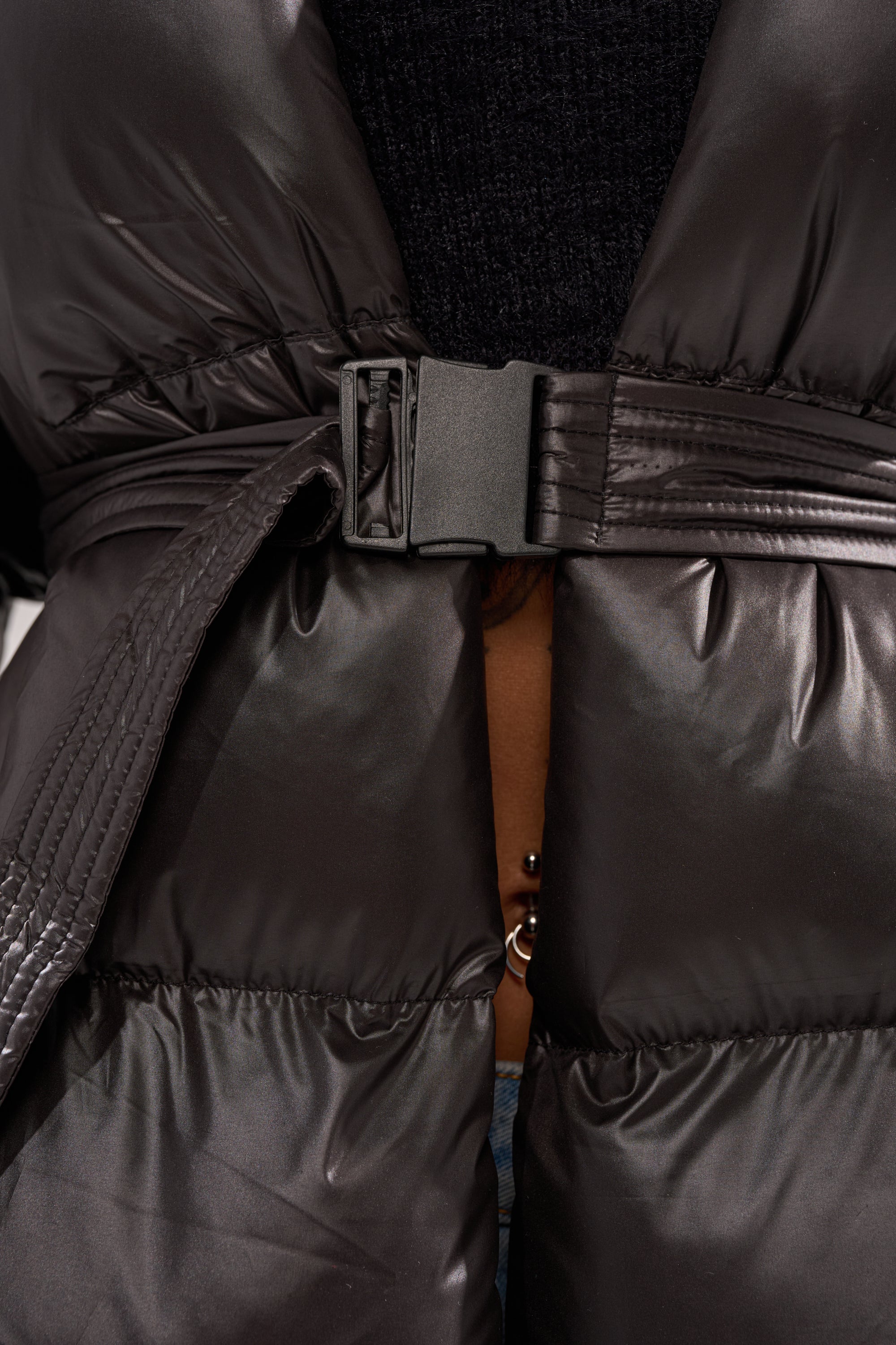 Close-up of the POPPIN OFF TEARAWAY PUFFER, a black belted puffer jacket fastened with a plastic buckle. The coat is partially open, showing a brown-skinned person's midriff and belly button piercing underneath.