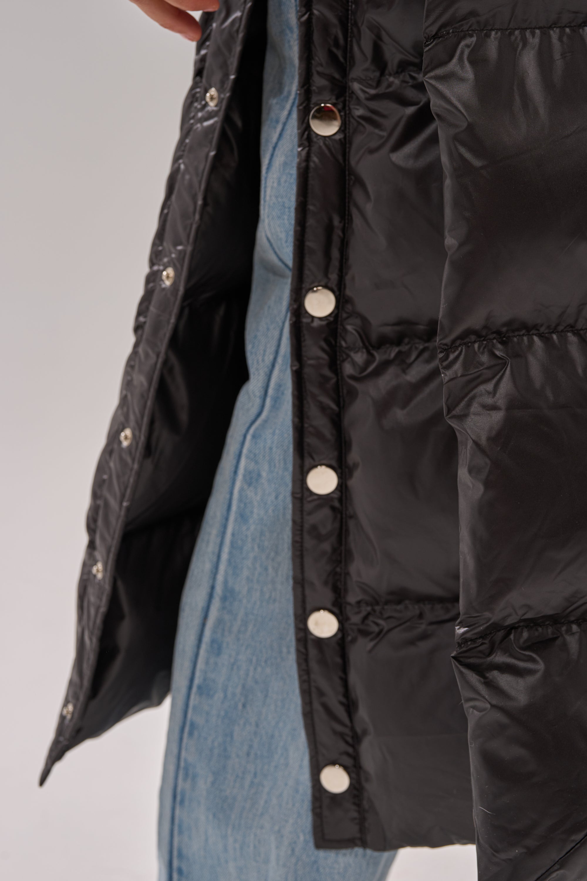 Close-up of a person wearing light blue jeans and the POPPIN OFF TEARAWAY PUFFER, a glossy black jacket with silver snap buttons, standing against a plain light background.