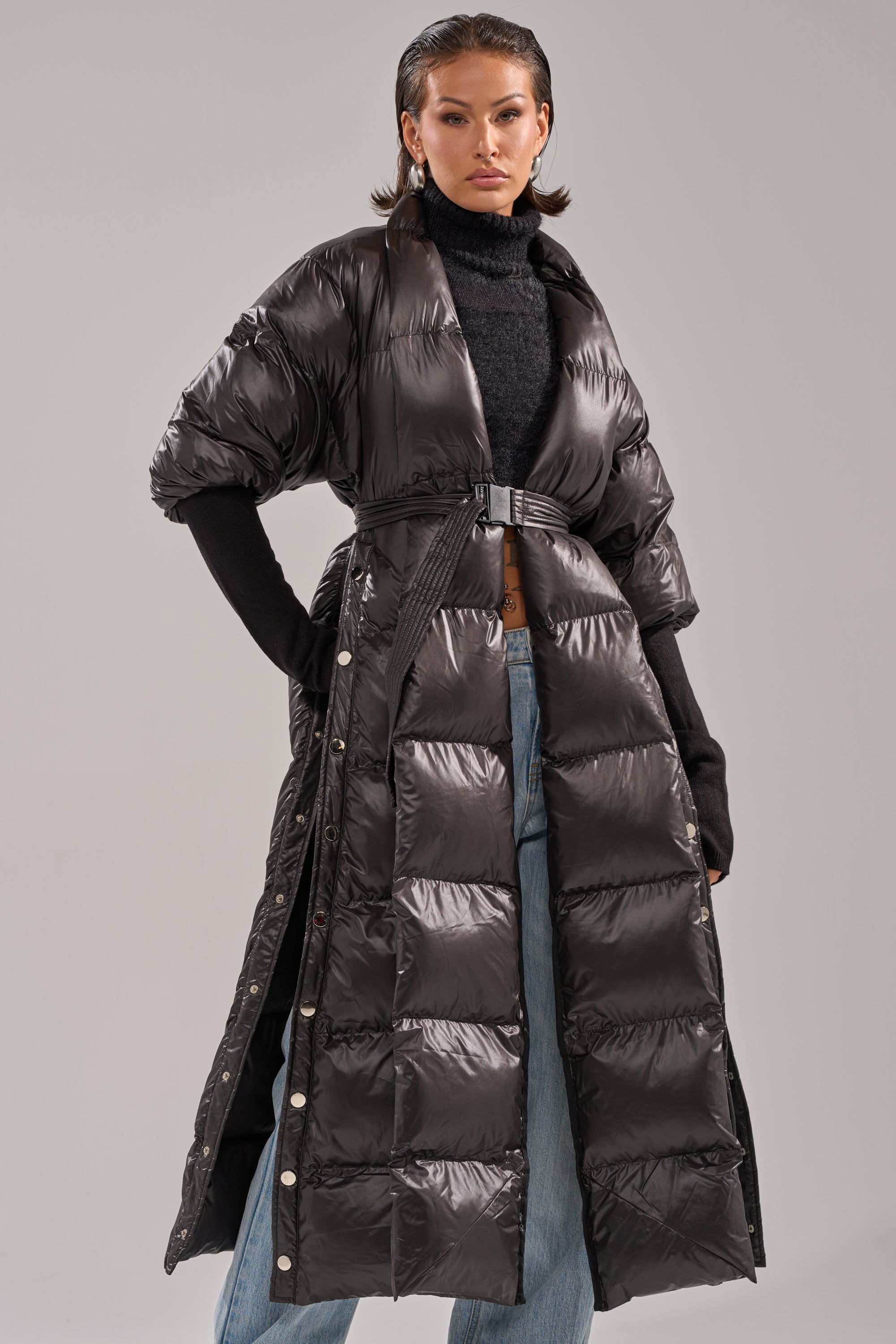 A person stands confidently against a plain light gray background wearing the POPPIN OFF TEARAWAY PUFFER—a glossy black belted short-sleeve puffer coat—paired with a black turtleneck and blue jeans.