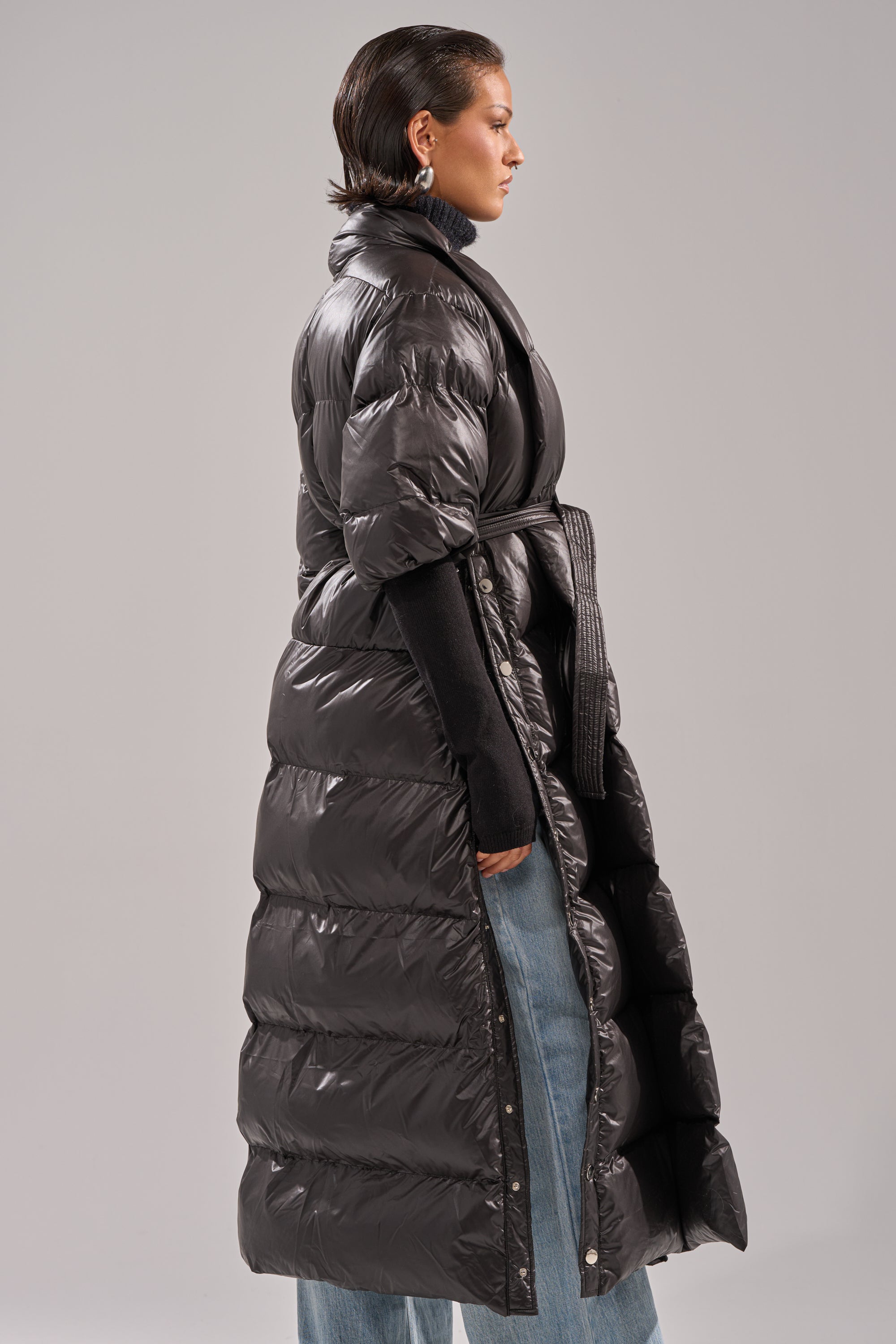 Wearing the POPPIN OFF TEARAWAY PUFFER, a glossy black short-sleeve puffer coat, over a black turtleneck and blue jeans, a person stands in profile against a plain grey background.