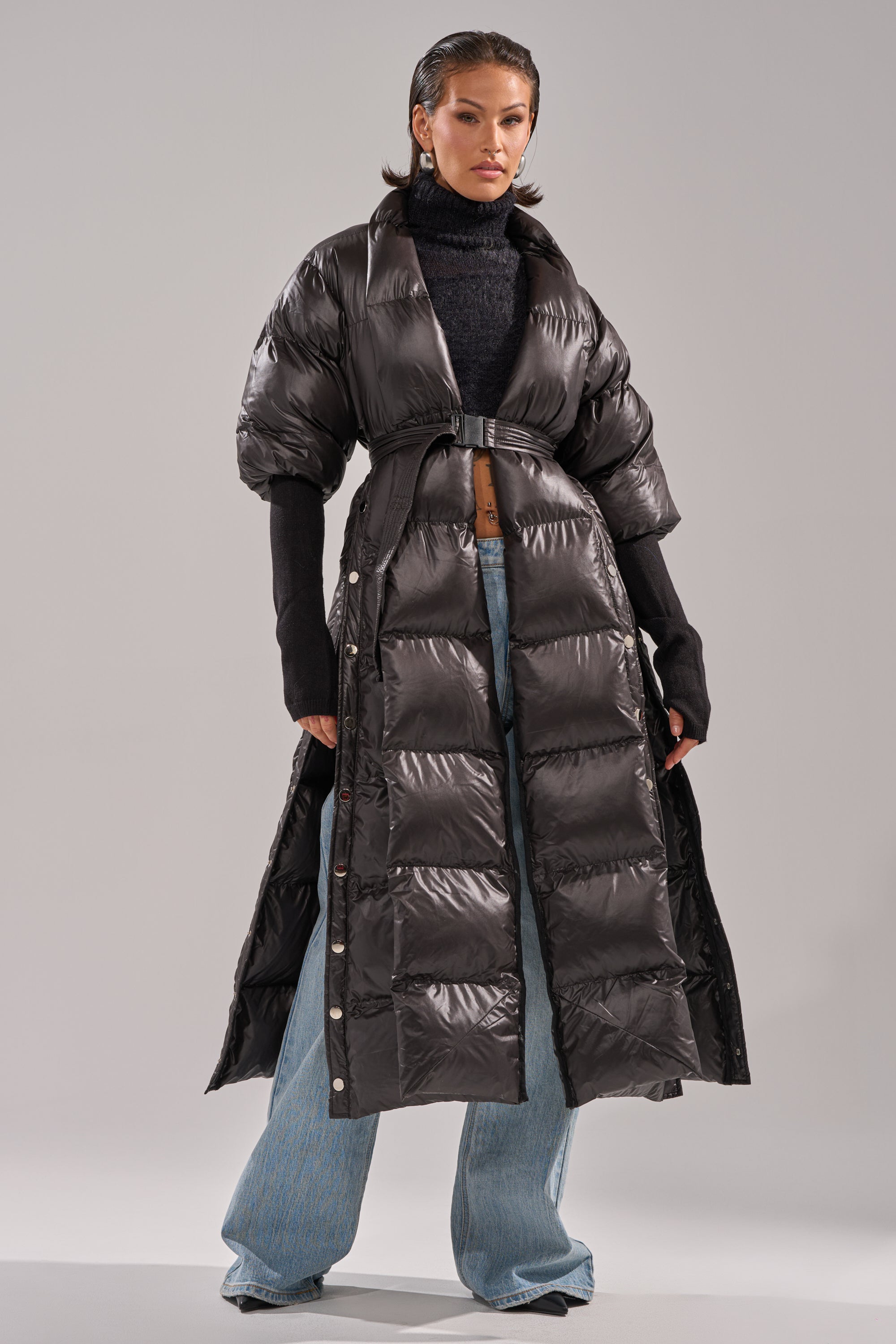 A person wearing the POPPIN OFF TEARAWAY PUFFER—a shiny black belted, short-sleeve puffer jacket—over a black turtleneck and wide-leg jeans, standing confidently against a plain light background.