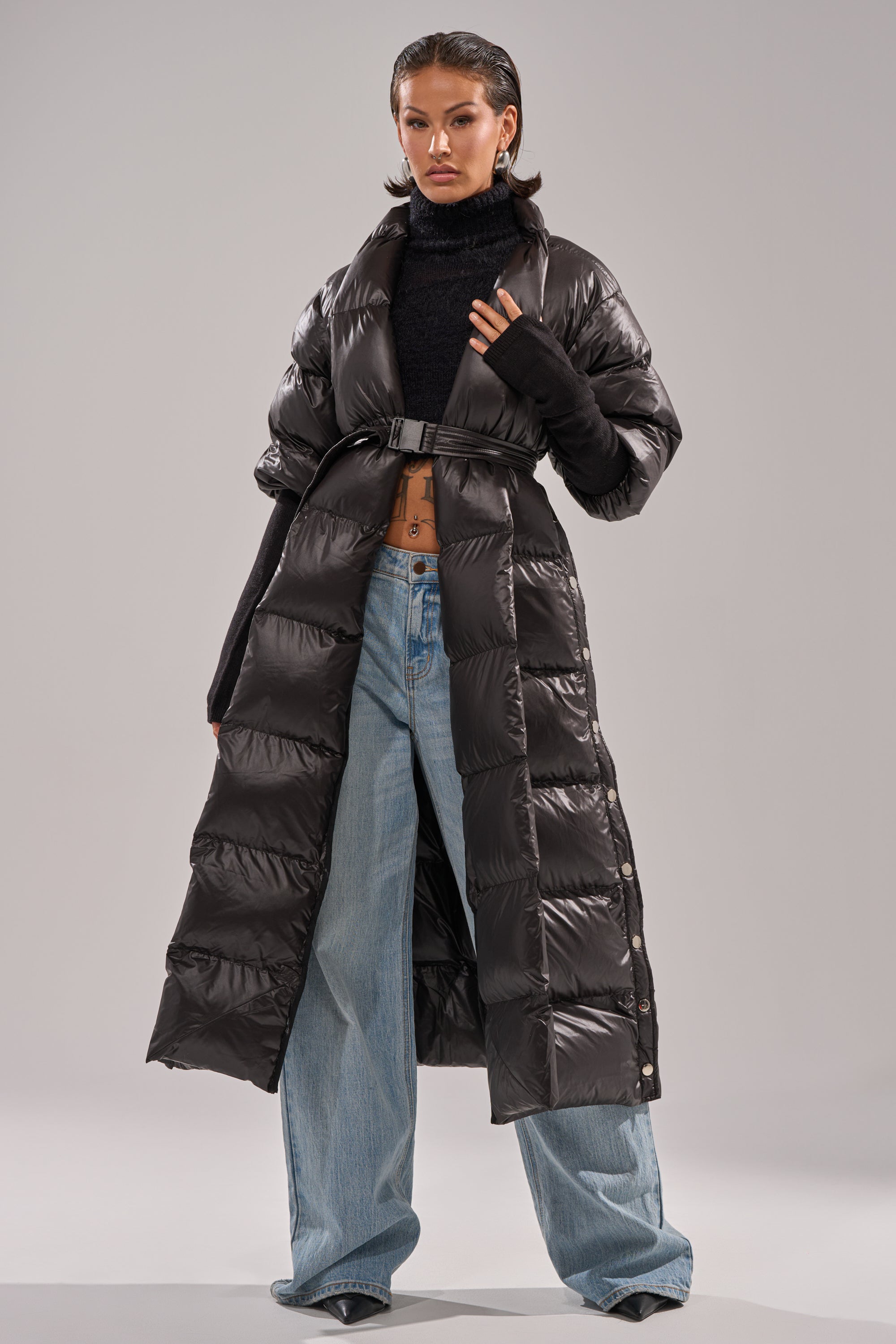 A woman poses confidently in the POPPIN OFF TEARAWAY PUFFER, styled over a black turtleneck crop top, wide-leg light blue jeans, and pointed black shoes. Her wet-look hair stands out against the plain gray background.