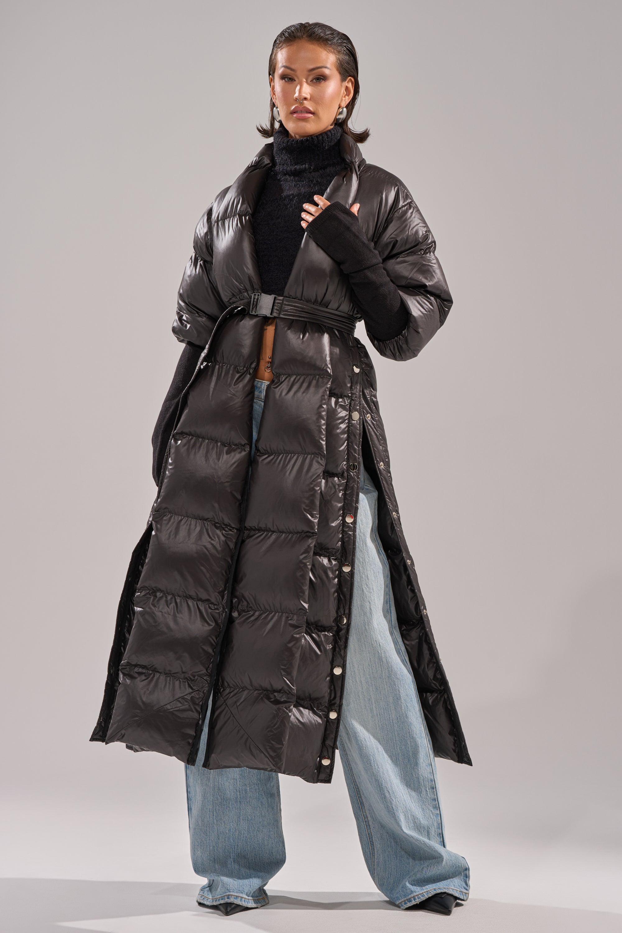A person stands confidently against a plain background, wearing the POPPIN OFF TEARAWAY PUFFER—a long black puffer coat with a belted waist—over a black turtleneck and wide-leg light blue jeans.