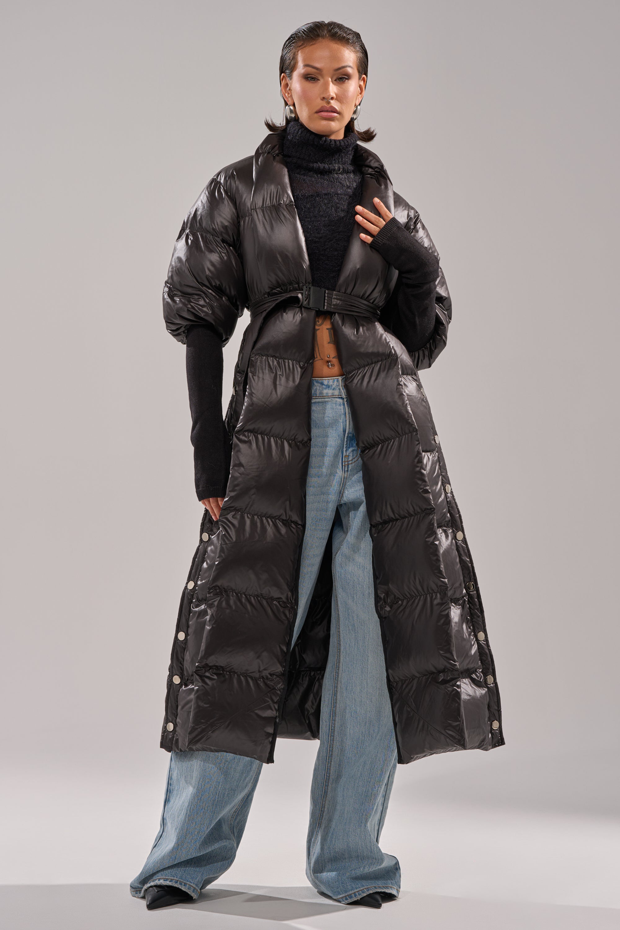 A woman wears the POPPIN OFF TEARAWAY PUFFER, a long black coat with a belted waist, styled over a black turtleneck and wide-leg blue jeans. Her hair is slicked back as she poses confidently against a light gray background.