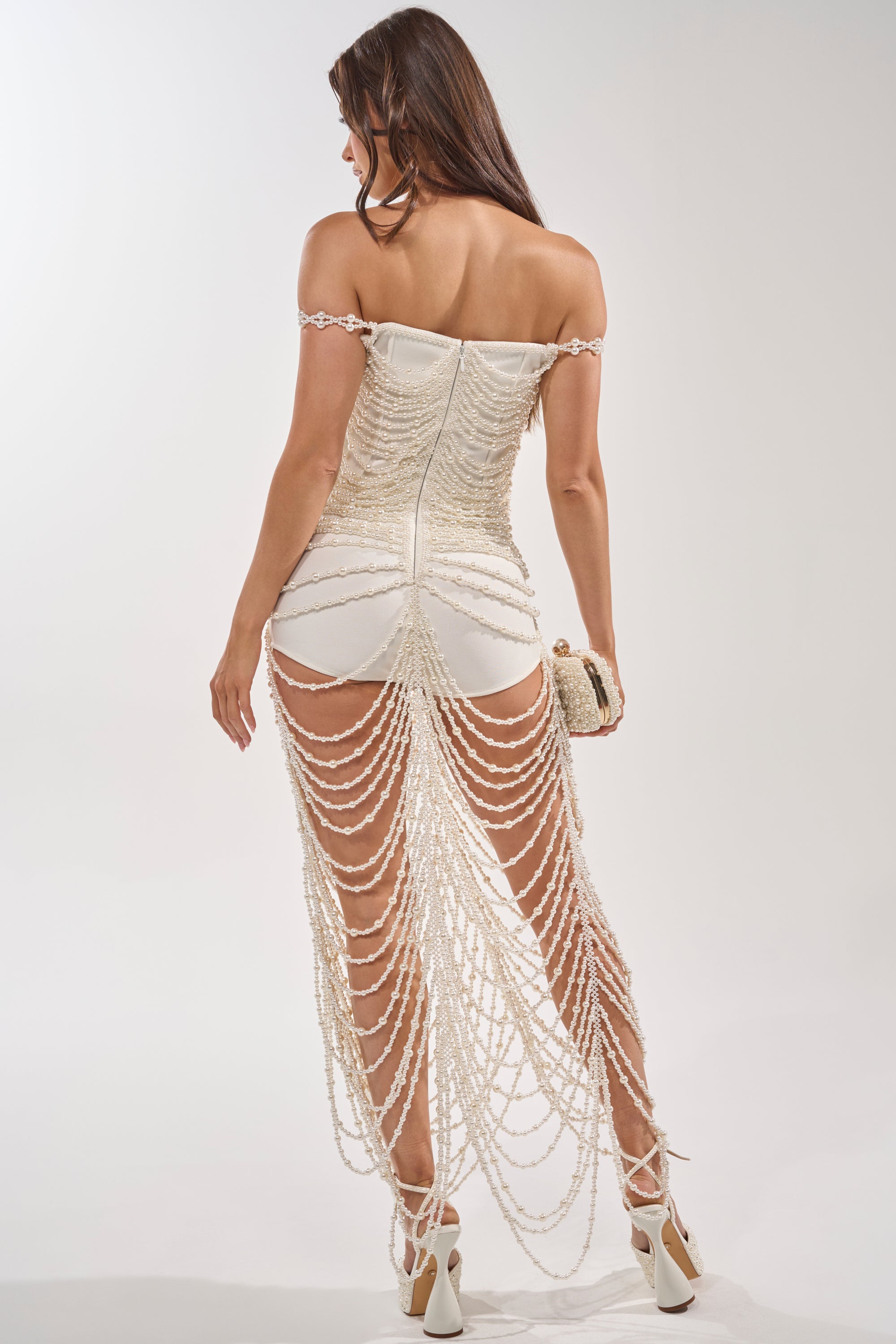 A woman faces away, wearing the DRESSED UP FOR THE BEST PEARL MAXI DRESS featuring pearl embellishments and cascading pearl chains—ideal for bridal celebrations or formal events. She holds a clutch and pairs the look with white lace-up high heels. 4