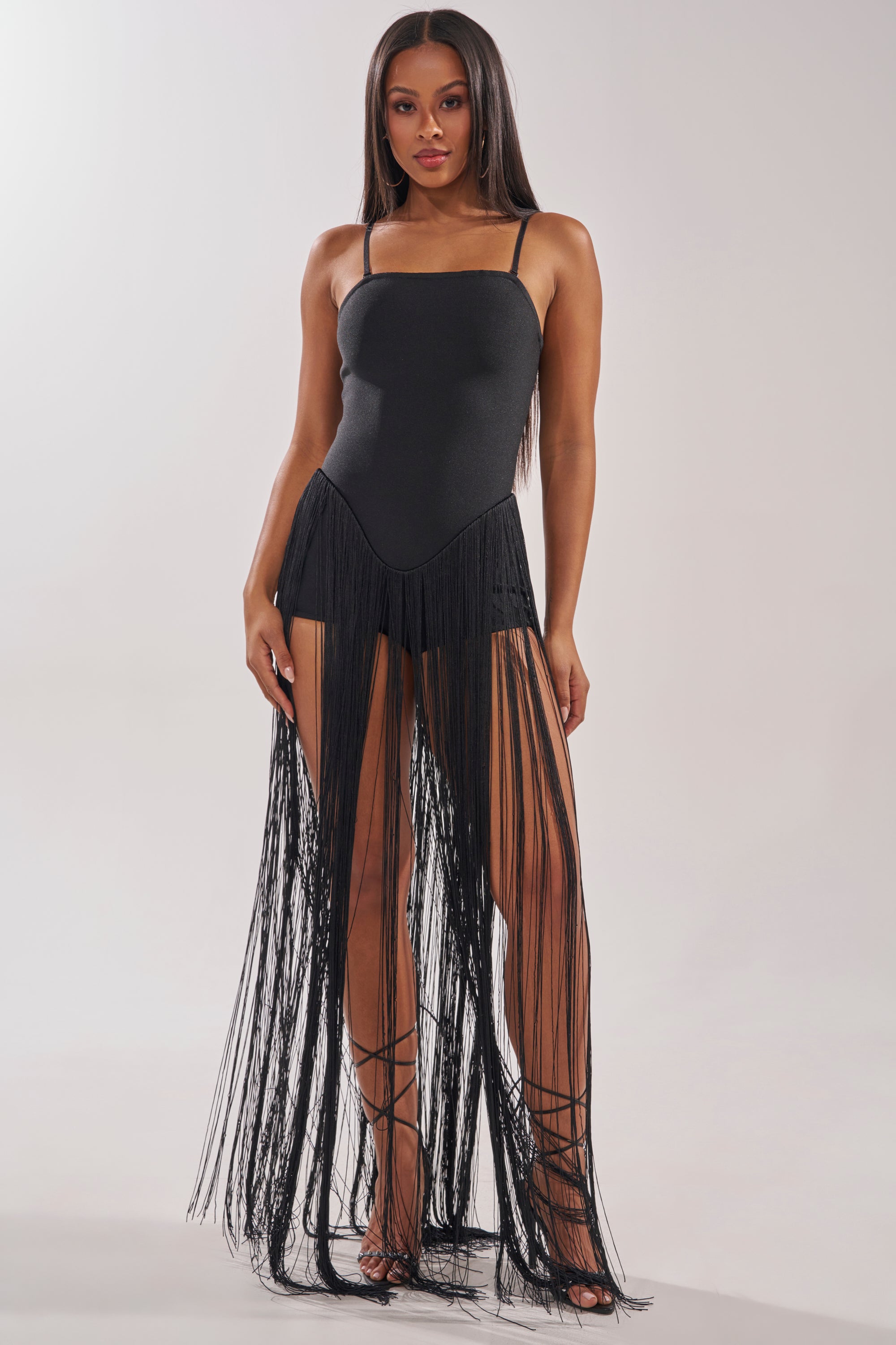 A woman faces forward in the TRY ME STRAPLESS FRINGE MAXI DRESS, featuring black fabric and floor-length fringe from the waist, styled with lace-up heels against a plain light background. 2