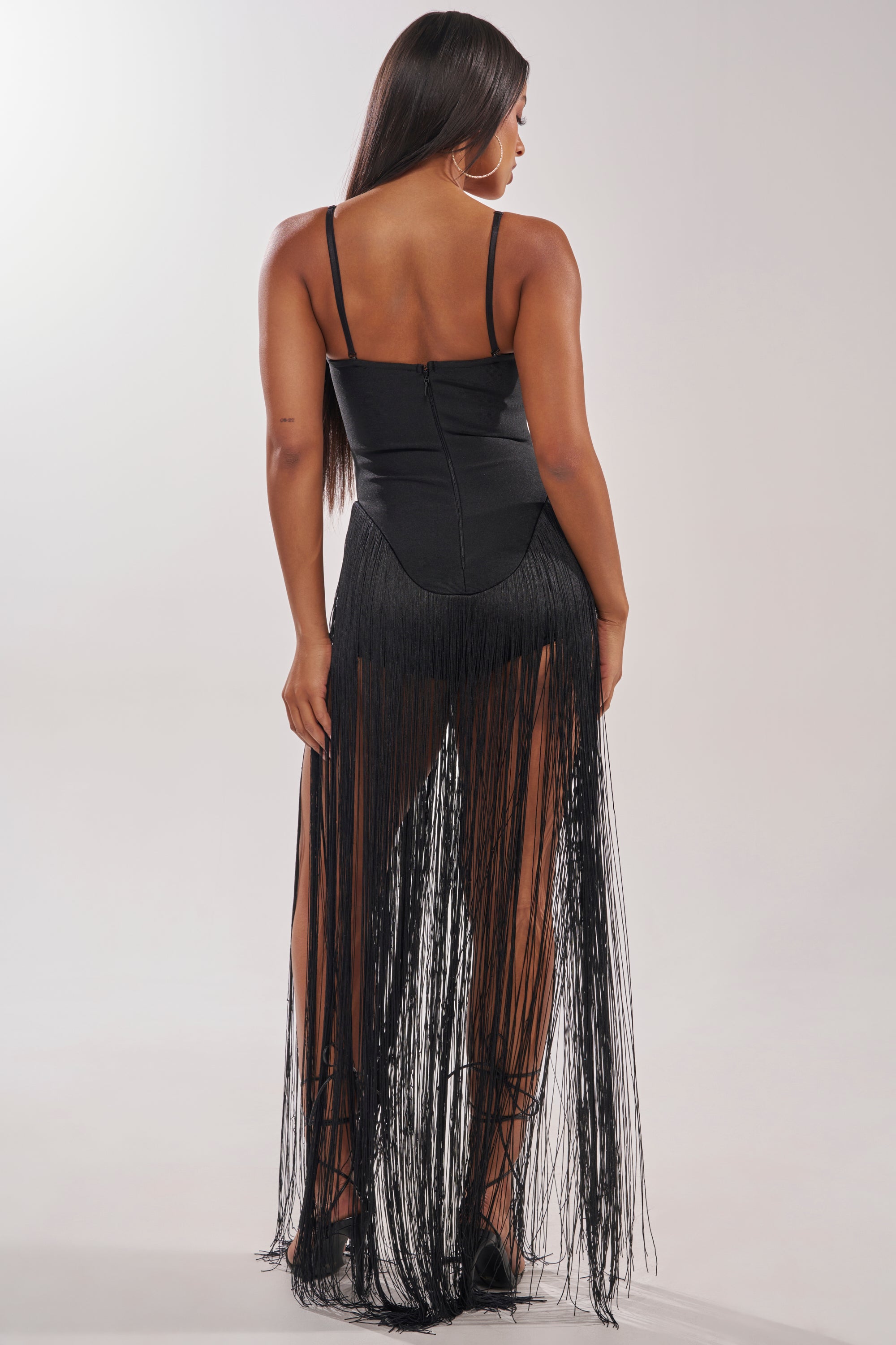 A woman faces away from the camera in the TRY ME STRAPLESS FRINGE MAXI DRESS, featuring black strapless styling and flowing sheer fringe from waist to floor—ideal for a chic nightlife look. She pairs it with long dark hair and hoop earrings. 4