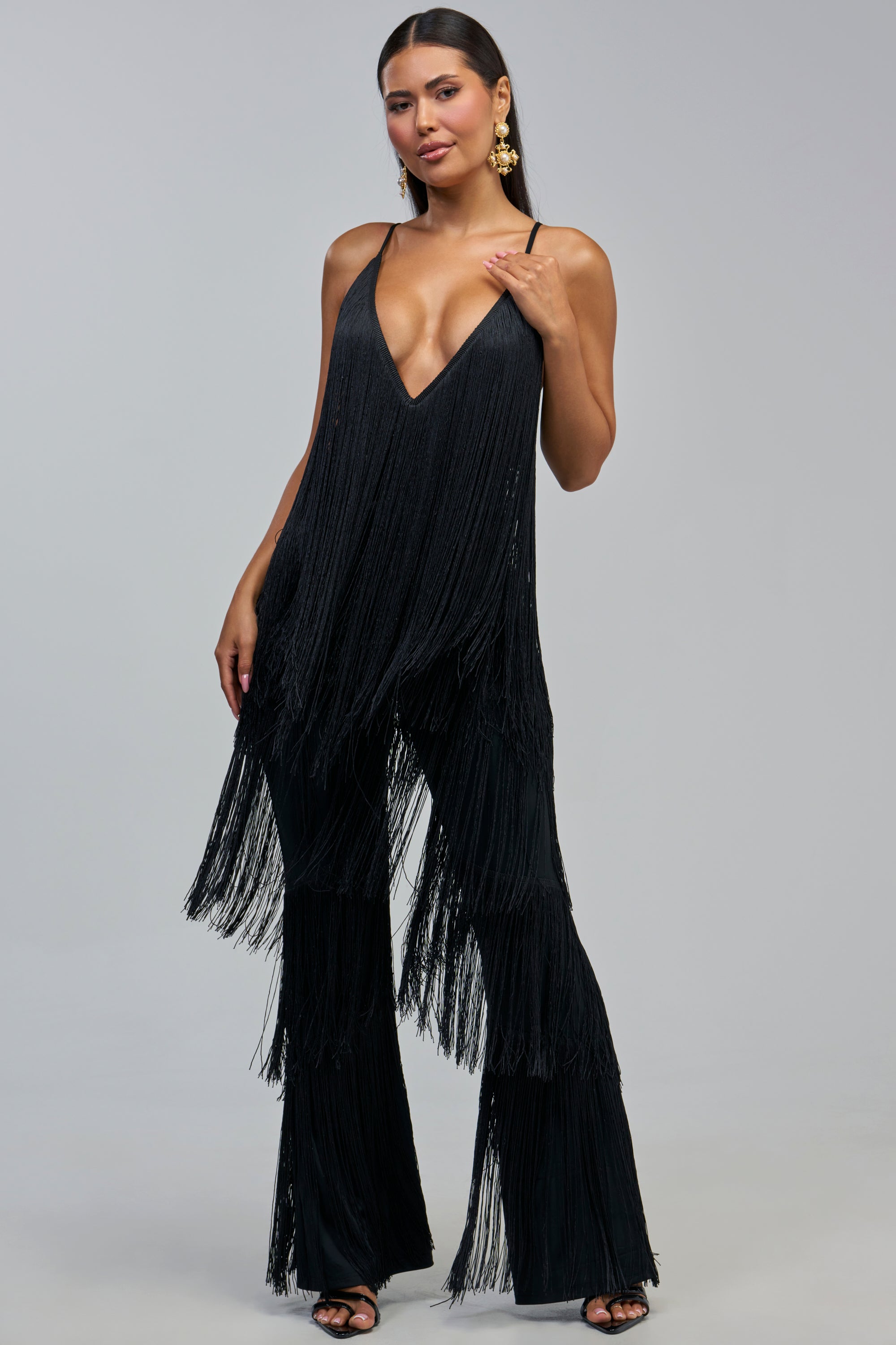 A woman poses in the BAECATION FRINGE JUMPSUIT IN BLACK, which features long fringe accents, a deep V-neckline, and wide-leg pants. She accessorizes with statement earrings and open-toe heels, her long hair styled back as she stands against a plain gray background—making this an ideal outfit for a girls’ night out.