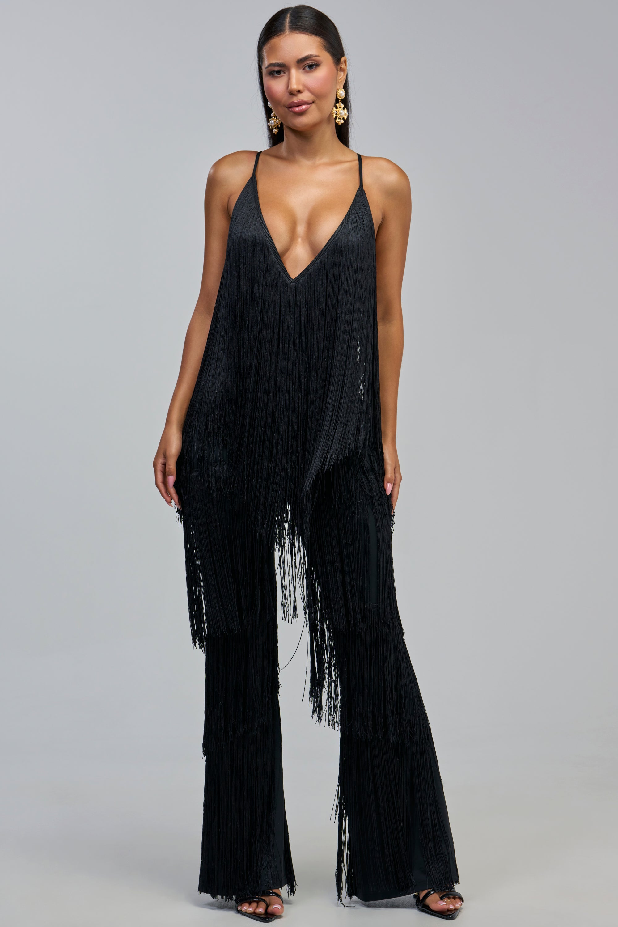 A woman wearing the BAECATION FRINGE JUMPSUIT IN BLACK—a sleeveless design featuring a deep V-neckline, wide-leg pants, and playful fringe—styled with statement earrings and black heels against a plain gray background; the perfect party jumpsuit for a chic girls’ night out.