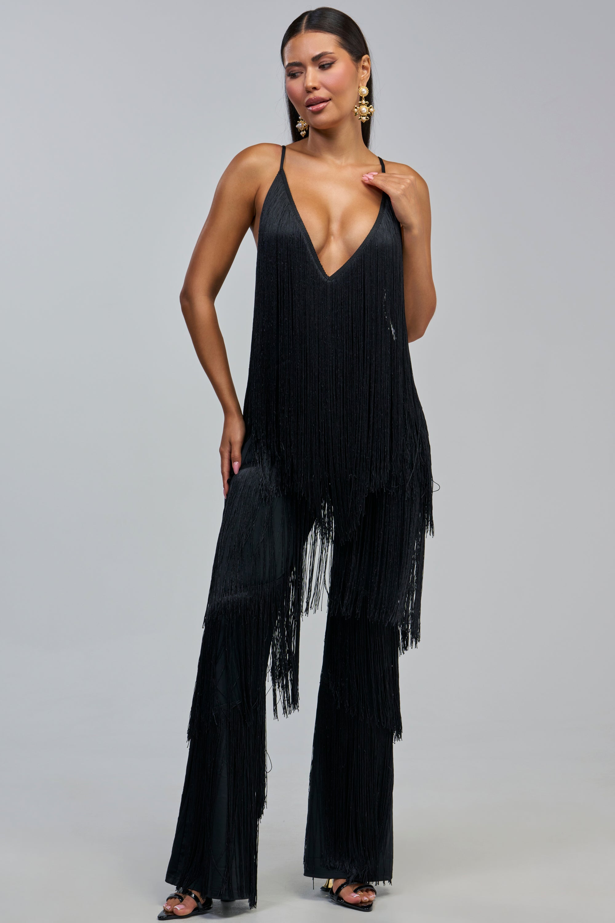 Model styled in the BAECATION FRINGE JUMPSUIT IN BLACK featuring a deep V-neckline and flared pants, paired with statement earrings and black heels—an ideal look for a girls’ night out, shot against a plain gray background.