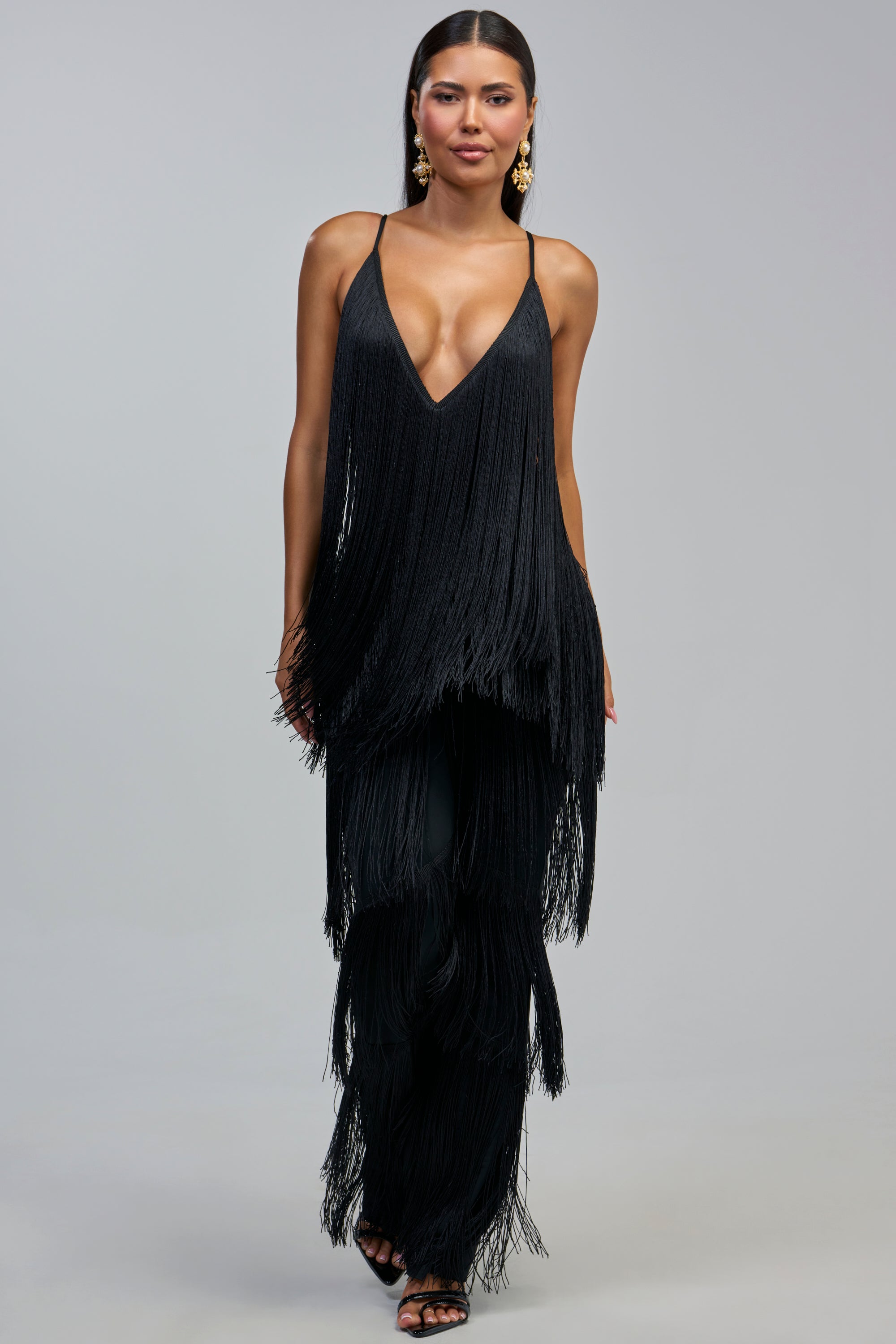A woman wears the BAECATION FRINGE JUMPSUIT IN BLACK—a sleeveless, deep V-neck black fringe jumpsuit—paired with gold earrings and black heels, standing against a light gray background. This is the perfect party look for your next girls’ night out.