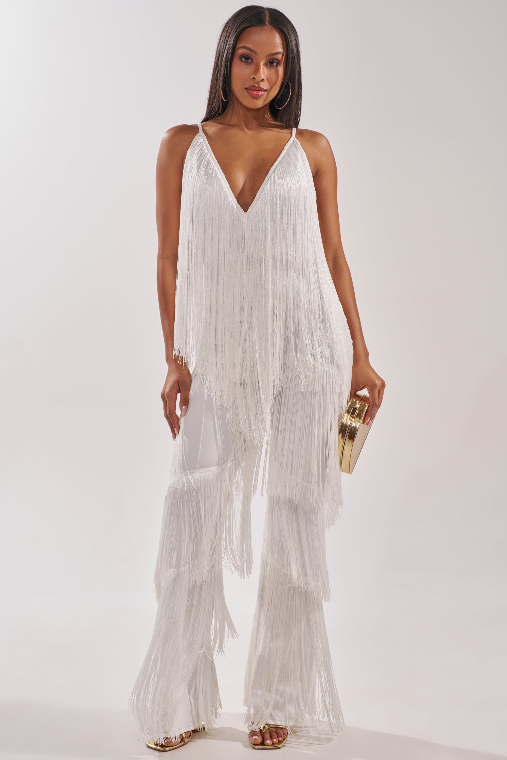 A woman poses against a plain light background in the BAECATION FRINGE JUMPSUIT—a party-ready white jumpsuit with a deep V-neckline, wide-leg pants, and fringed details—holding a gold clutch. 0
