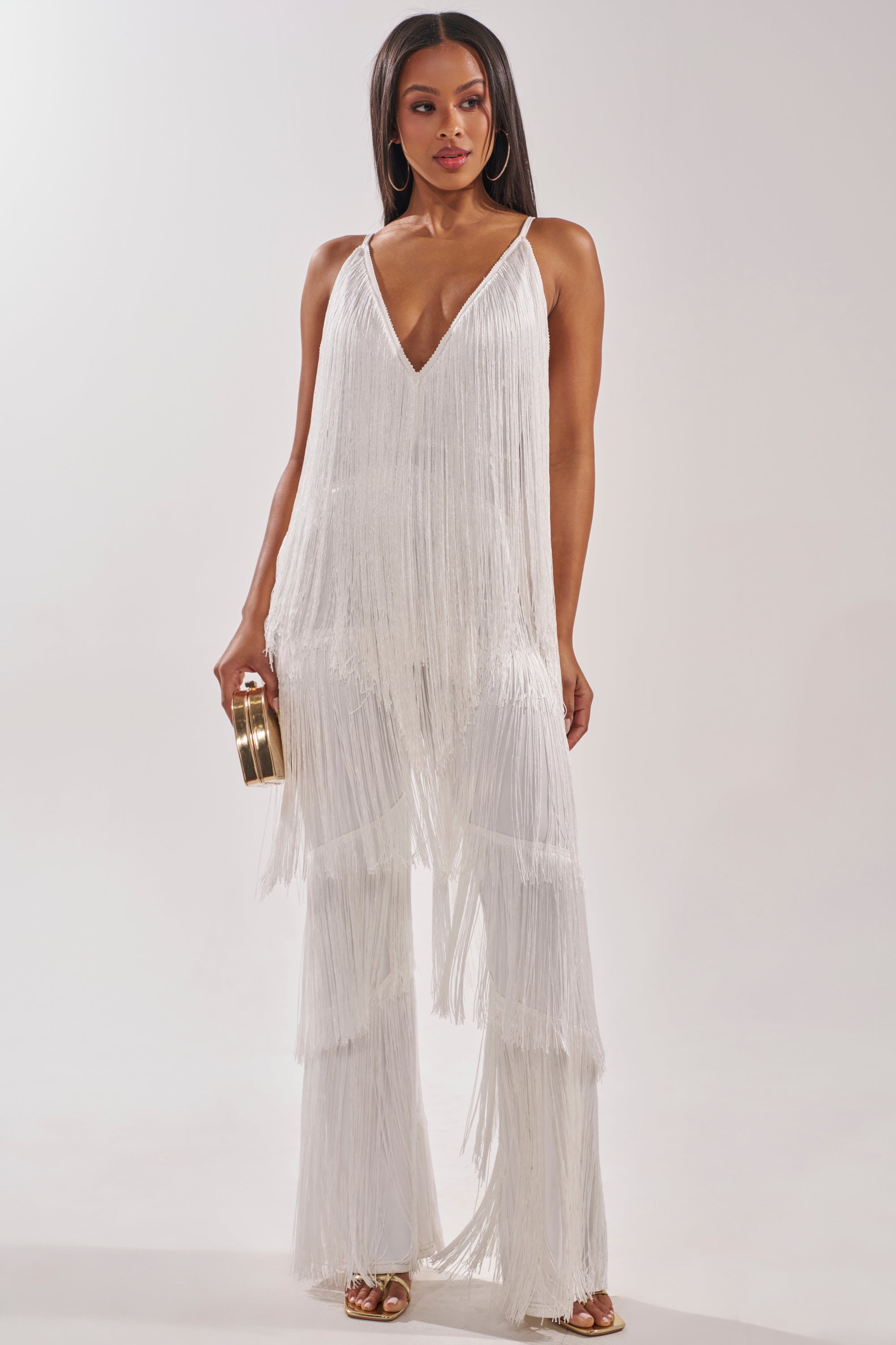 A woman wears the BAECATION FRINGE JUMPSUIT featuring a sleeveless design, white fringe details, deep V-neckline, and wide-leg pants. She holds a gold clutch and stands in gold high-heeled sandals against a light background. 1