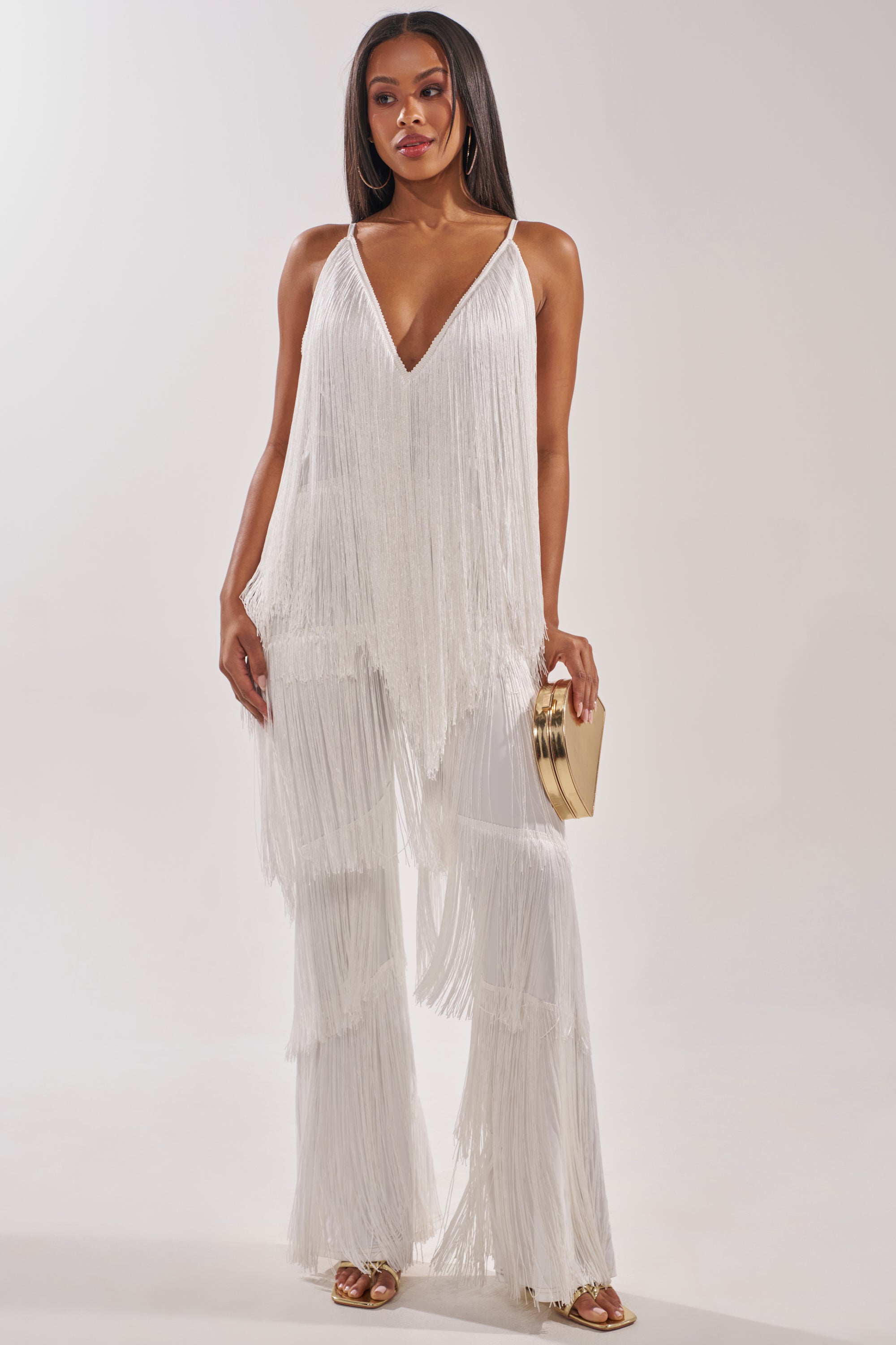 A woman models the BAECATION FRINGE JUMPSUIT, featuring wide-leg pants and white fringe, accessorized with a gold clutch and gold sandals, posed against a plain light background. 2
