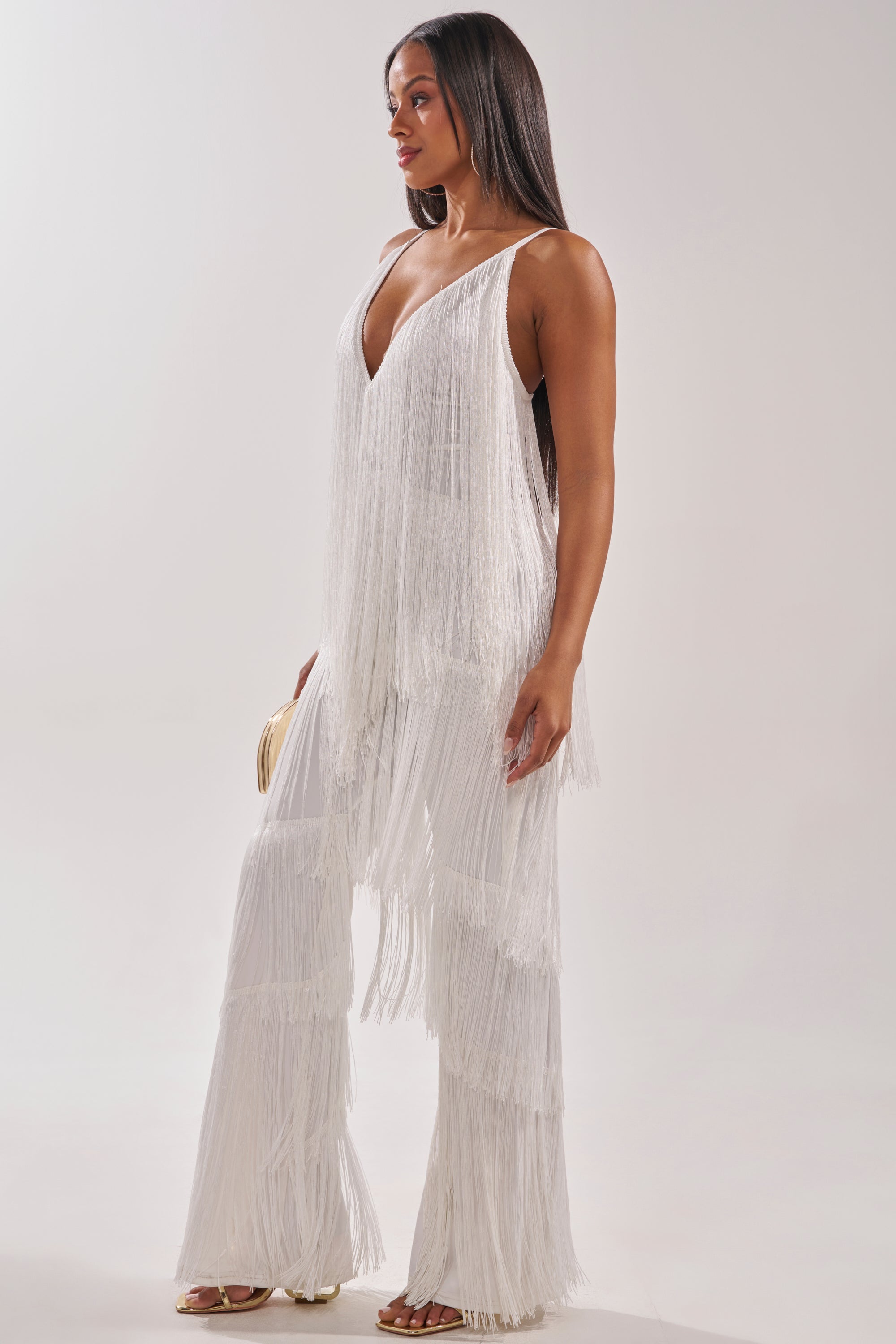 A woman stands in profile wearing the BAECATION FRINGE JUMPSUIT, a white party-ready piece with wide-leg pants, a deep V-neck, and tiered fringe. She pairs it with open-toed heels and a beige clutch against a plain light background. 3