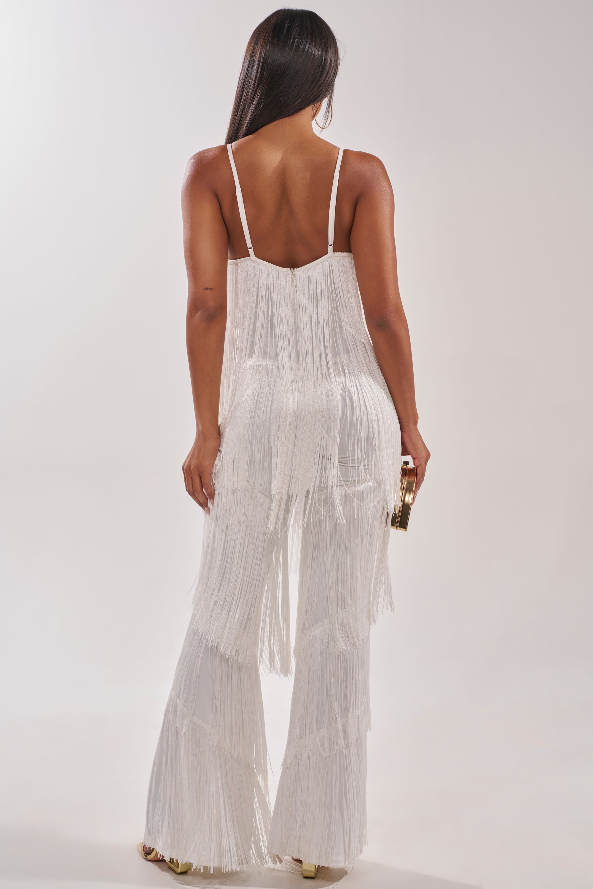 A woman with long dark hair, seen from the back, wears the BAECATION FRINGE JUMPSUIT featuring wide-leg pants and thin straps. She holds a small gold clutch and stands against a plain light background. 4