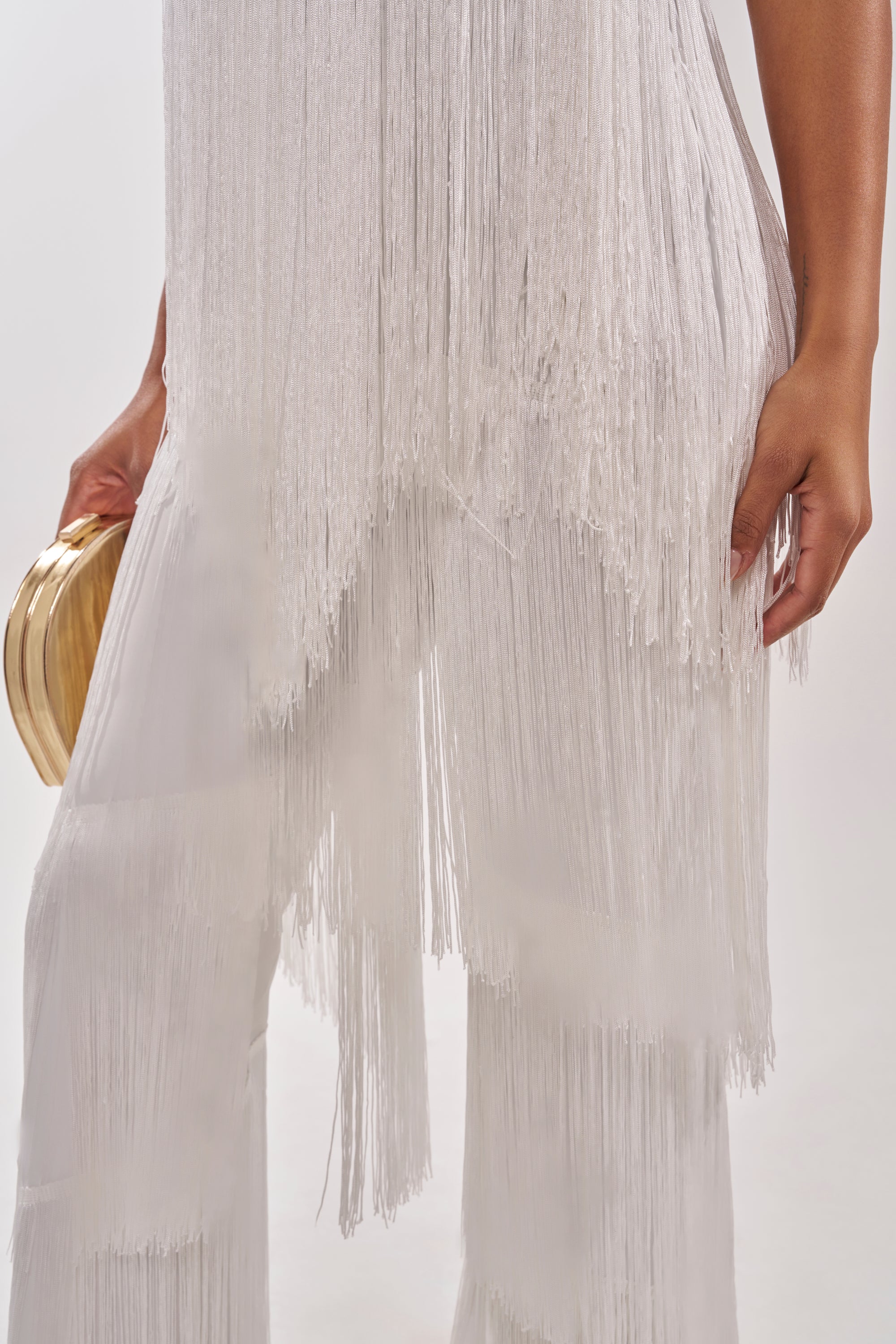 A person models the BAECATION FRINGE JUMPSUIT in white, holding a small gold clutch. The photo highlights the wide-leg silhouette and flowing fringe details on the lower torso and upper legs for a festive look. 5