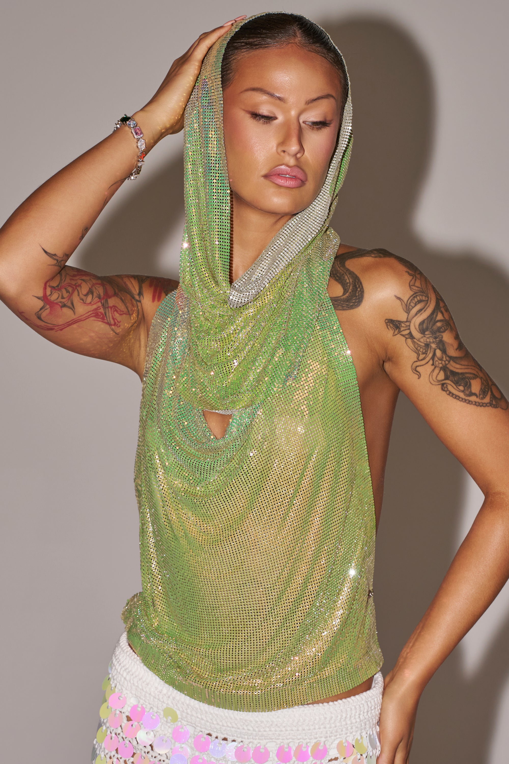 A person poses with one arm raised, showing tattoos on both arms while wearing the IN MY MIND HOODED CHAINMAIL TOP IN GREEN and a white sequin skirt, standing against a plain background. 0