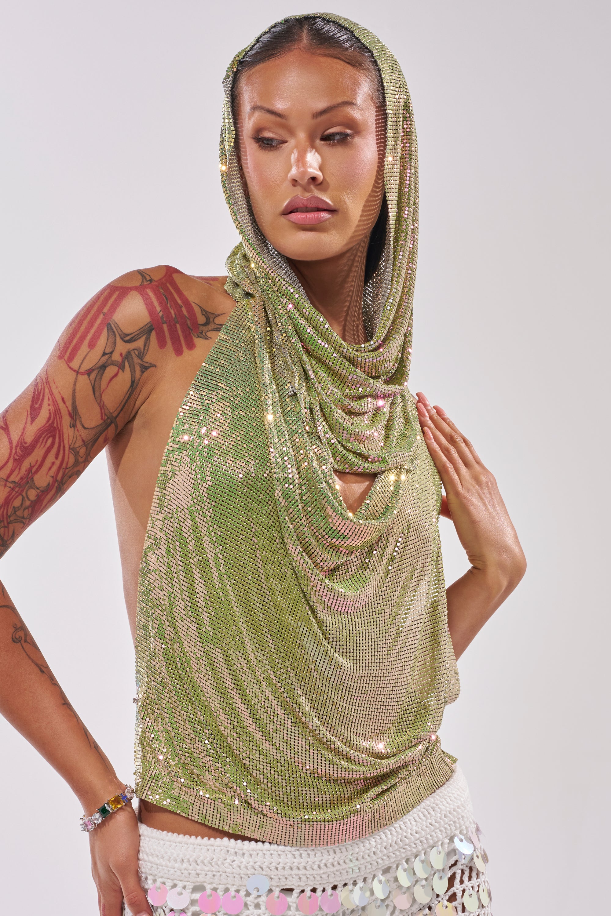 A person poses confidently in the IN MY MIND HOODED CHAINMAIL TOP IN GREEN and a white skirt with heart accents, showcasing arm tattoos against a plain, light background. 1