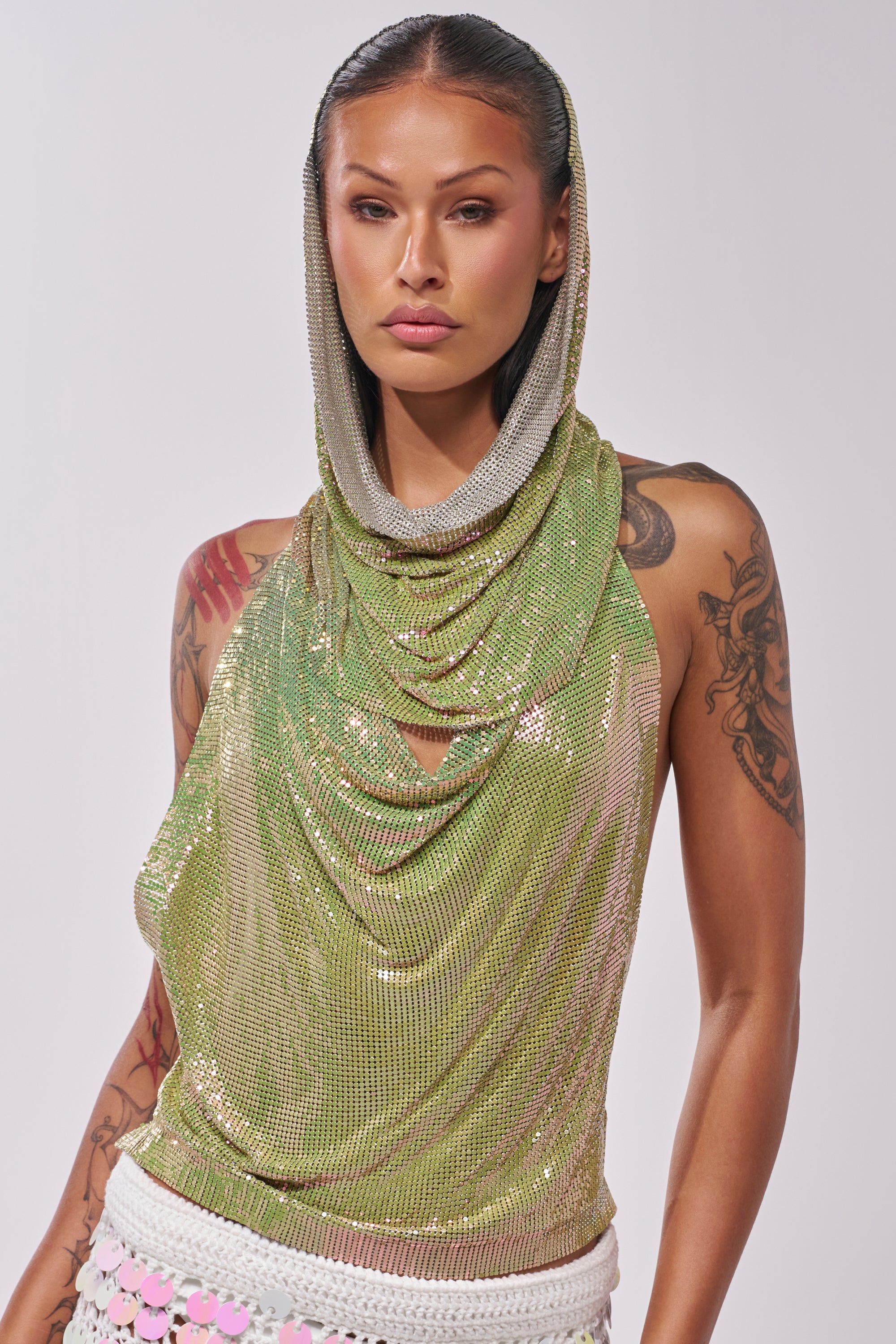 A woman with long, slicked-back hair and tattoos wears the IN MY MIND HOODED CHAINMAIL TOP IN GREEN with a white crocheted skirt, standing confidently against a plain light background. 2