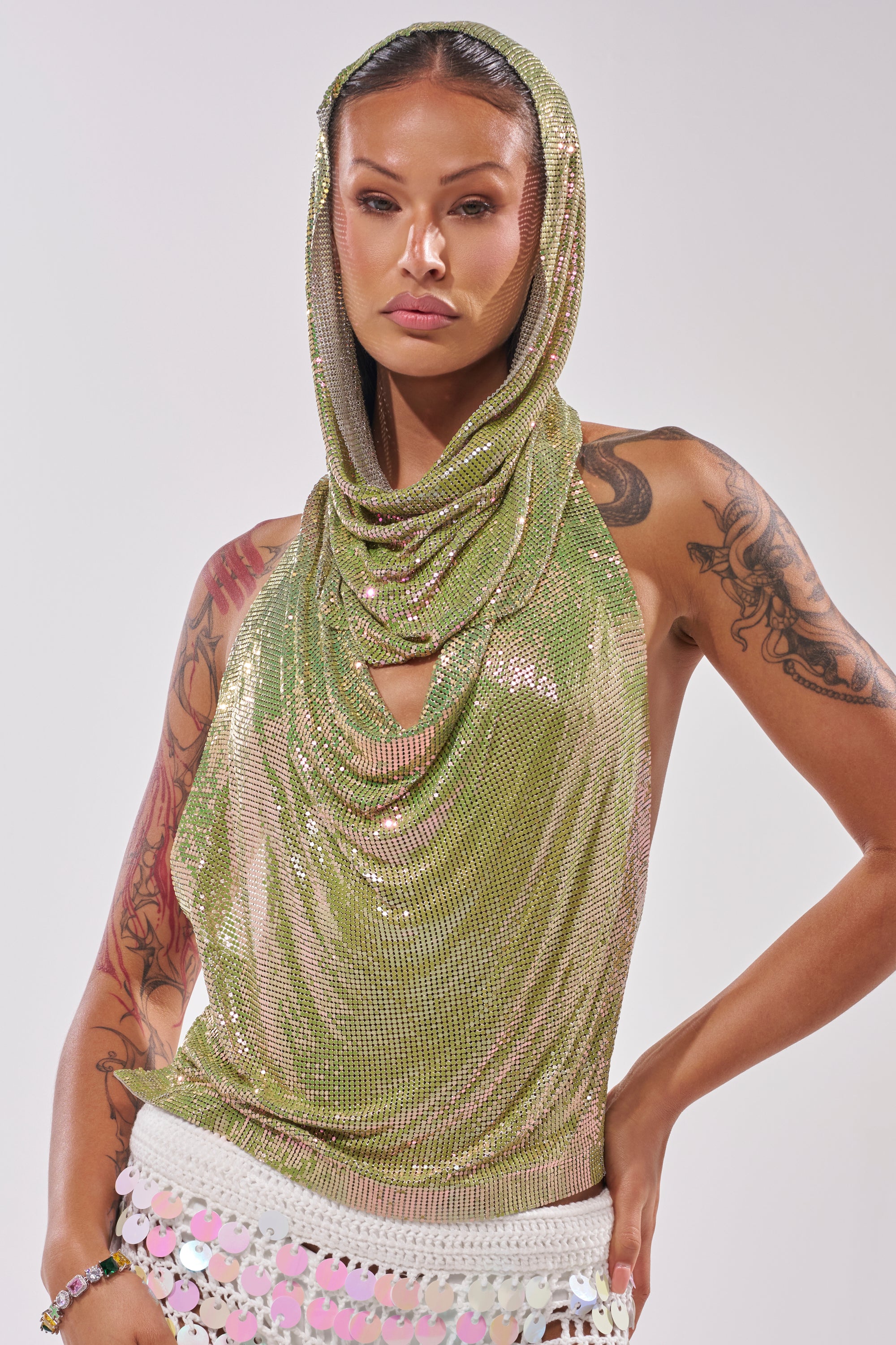 A person stands confidently in the IN MY MIND HOODED CHAINMAIL TOP IN GREEN with a draped neckline, visible tattoos on their arms, and a white skirt adorned with decorative circular accents for a striking runway partywear look. 3