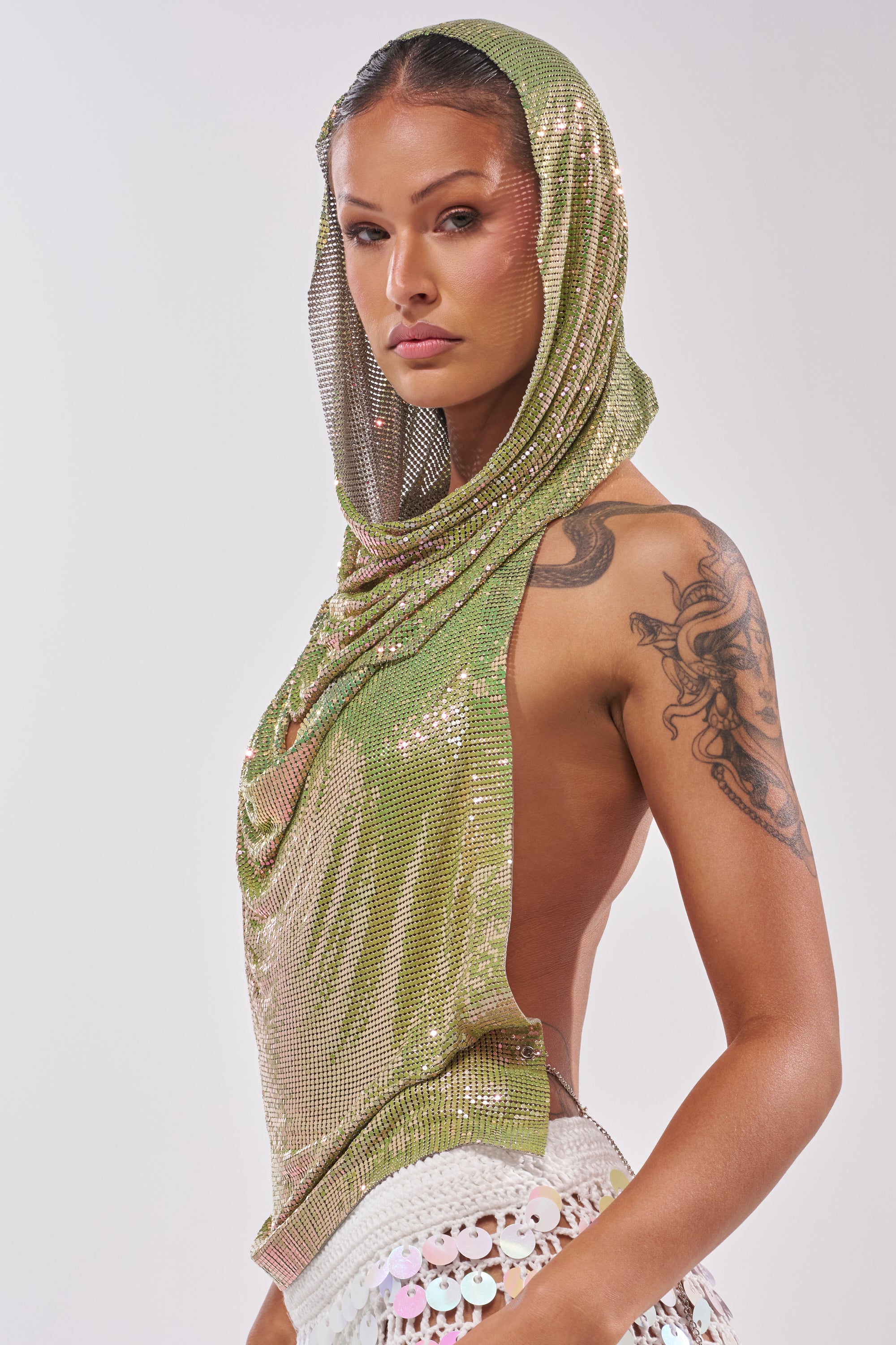 A woman with a visible arm tattoo wears the IN MY MIND HOODED CHAINMAIL TOP IN GREEN and a white sequin skirt, posing confidently against a light background—a bold, runway-ready party look. 4