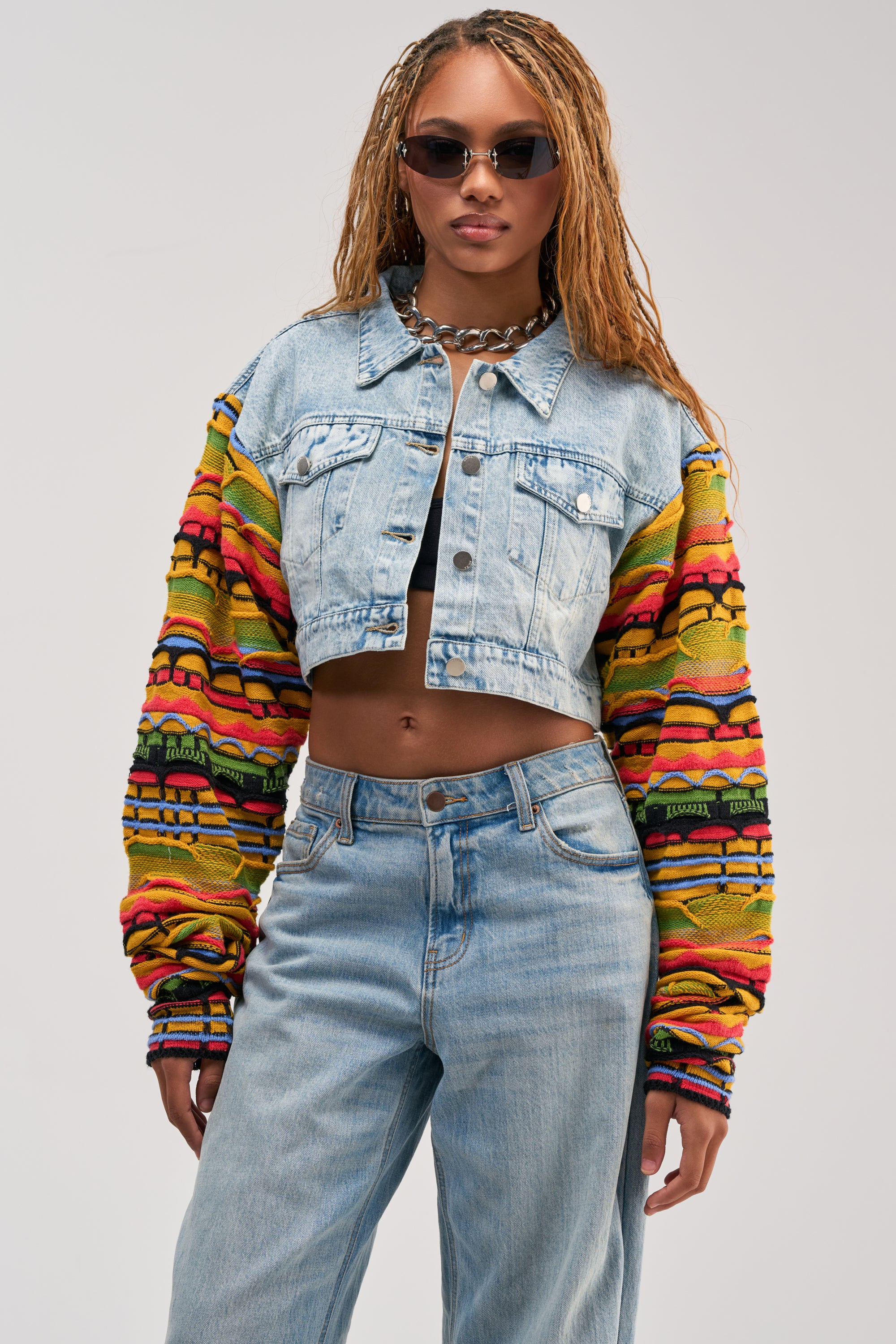 Woman wearing sunglasses, a black crop top, layered necklaces, light blue jeans, and the THROWBACK KNIT SLEEVE DENIM JACKET—featuring a retro-chic cropped fit with colorful knit sleeves—standing against a plain background.