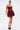 A woman stands confidently in the BELINA LACE MINI DRESS WITH BUBBLE SKIRT IN BURGUNDY, featuring a strapless sheer bodice and voluminous skirt. She holds a gold clutch, wears platform heels, and her hair is slicked back against a white background.