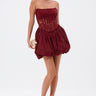 A woman stands confidently in the BELINA LACE MINI DRESS WITH BUBBLE SKIRT IN BURGUNDY, featuring a strapless sheer bodice and voluminous skirt. She holds a gold clutch, wears platform heels, and her hair is slicked back against a white background.
