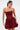 A woman wears the BELINA LACE MINI DRESS WITH BUBBLE SKIRT IN BURGUNDY, paired with a small gold clutch. She styles her hair in a sleek ponytail and accessorizes with hoop earrings.