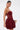 A woman with dark hair in a sleek ponytail wears the BELINA Lace Mini Dress with Bubble Skirt in Burgundy, featuring a strapless lace bodice and voluminous skirt. She holds a gold clutch against a plain light background.