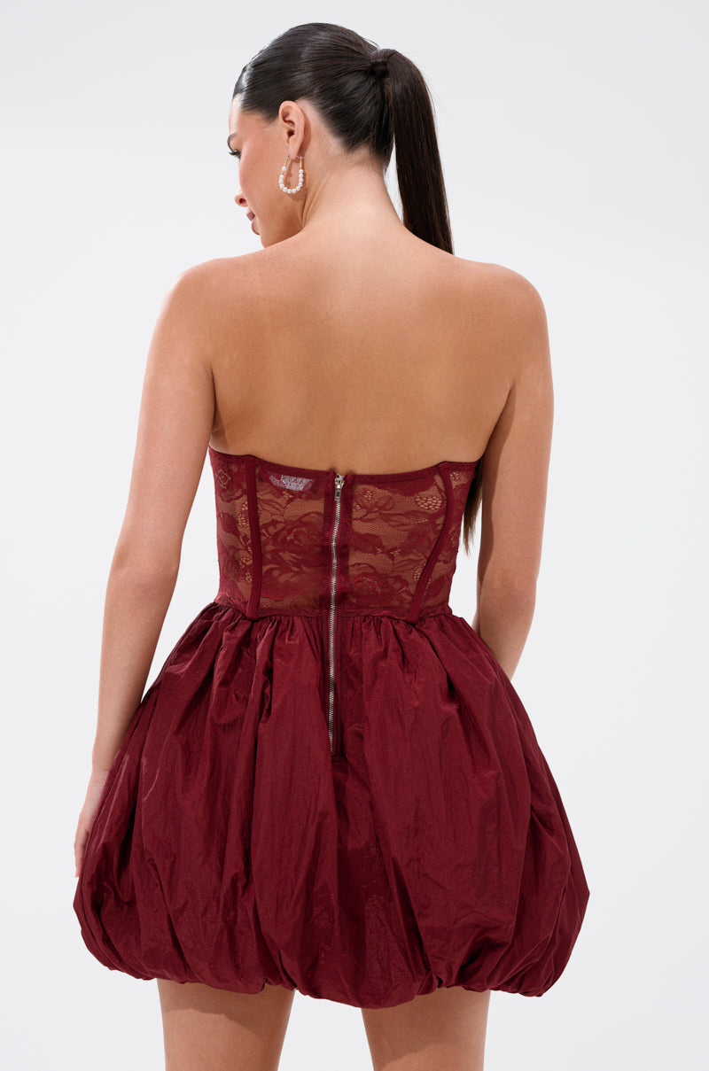 A woman with long dark hair in a ponytail wears the BELINA LACE MINI DRESS WITH BUBBLE SKIRT IN BURGUNDY, which features a corset back and bubble skirt, standing against a plain light background.