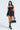 A woman in the BELINA LACE MINI DRESS WITH BUBBLE SKIRT strides confidently. She complements her strapless black dress, featuring a sheer corset and sweetheart neckline, with a black handbag and knee-high lace-up boots. Her long hair flows freely, adorned with hoop earrings.
