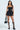 A woman models the BELINA LACE MINI DRESS, which is strapless with a sweetheart neckline and sheer corset top adorned with lace. She accessorizes with a black bag, knee-high buckle boots, and hoop earrings as her long dark hair gracefully cascades.