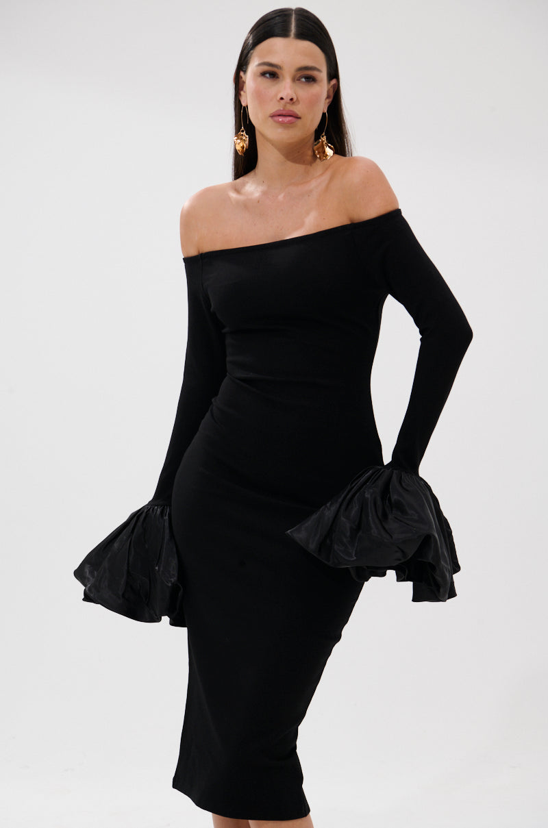 A woman models the CLASS WITH A LITTLE BIT OF SASS MIDI DRESS—an off-the-shoulder, fitted cocktail dress with long sleeves and ruffled cuffs. She pairs it with gold statement earrings and stands against a simple light background.