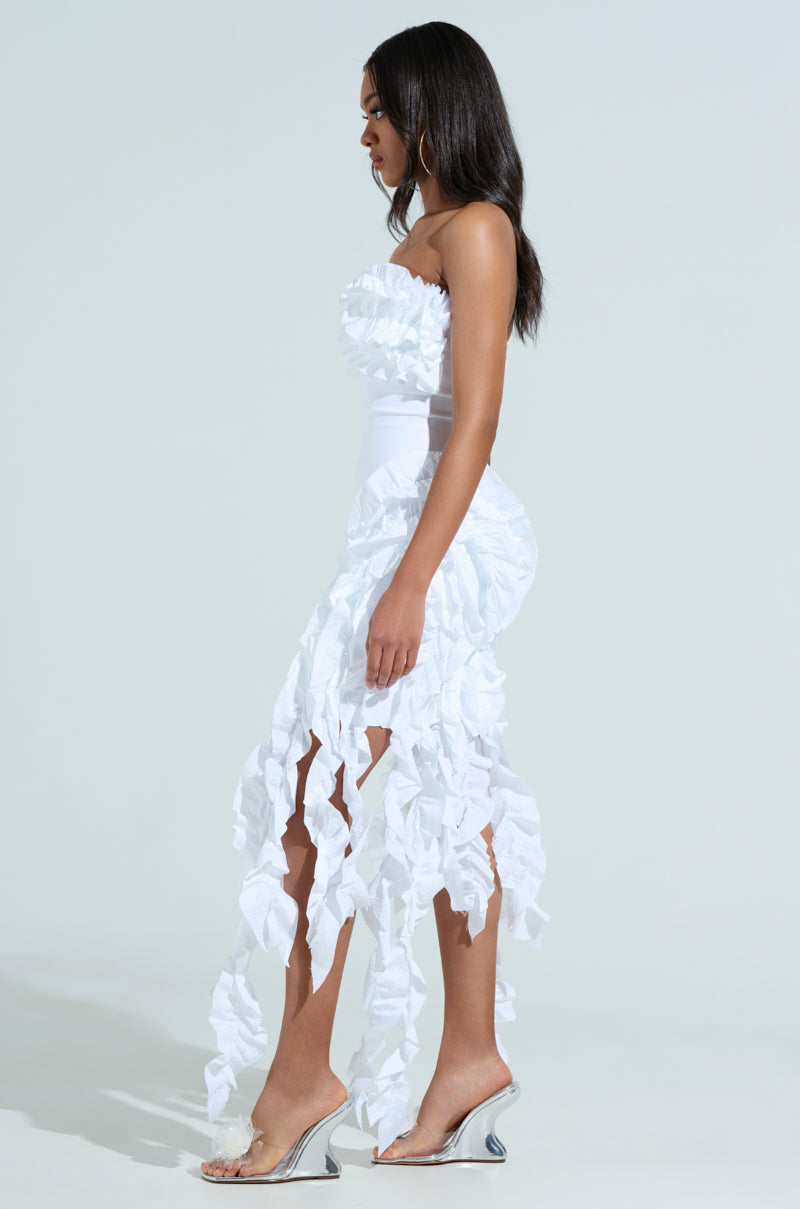 A woman stands sideways in the City Lights Ruffle Midi Dress in white, featuring textured fabric and a flowing cut-out skirt. Her long dark hair cascades like ruffled tendrils as she wears clear high-heeled shoes against a plain background.