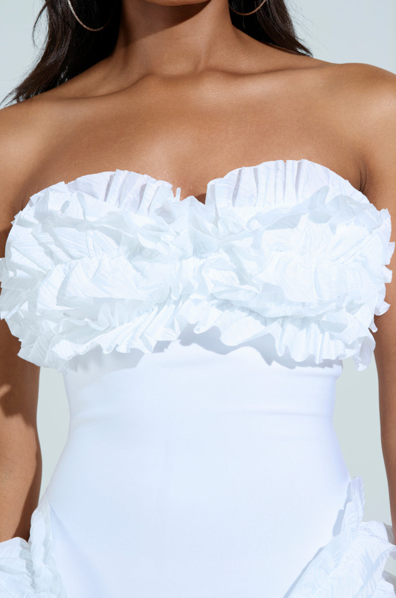 A woman poses in the City Lights Ruffle Midi Dress in white, featuring a textured design around the bust with intricate ruffled tendrils. The background is plain and light gray, highlighting the dress's details.