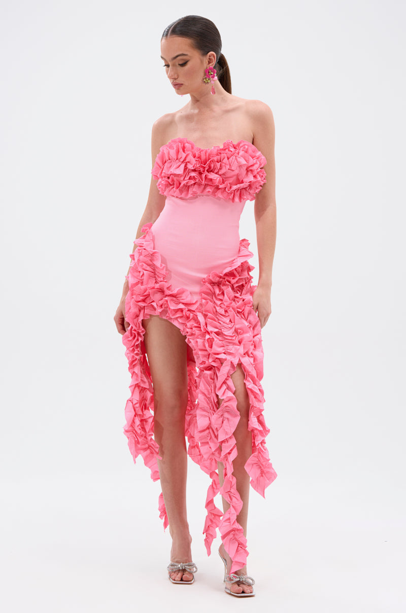 A woman stands against a white background wearing the CITY LIGHTS RUFFLE MIDI DRESS IN LIGHT PINK, a strapless, fitted dress with ruffled bodice details and an asymmetrical skirt, paired with silver heels and pink statement earrings.
