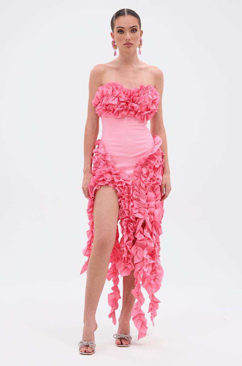 A woman confidently models the CITY LIGHTS RUFFLE MIDI DRESS IN LIGHT PINK, a strapless, form-fitting dress with dramatic ruffles and a high slit, styled with silver heels and statement earrings against a plain white background.