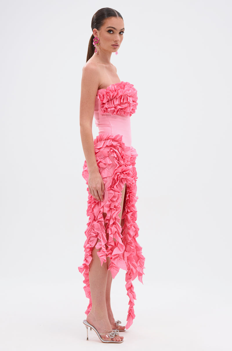 A woman poses against a white background wearing the CITY LIGHTS RUFFLE MIDI DRESS IN LIGHT PINK—strapless, fitted, with ruffled details and a high slit—paired with silver heels and pink statement earrings.