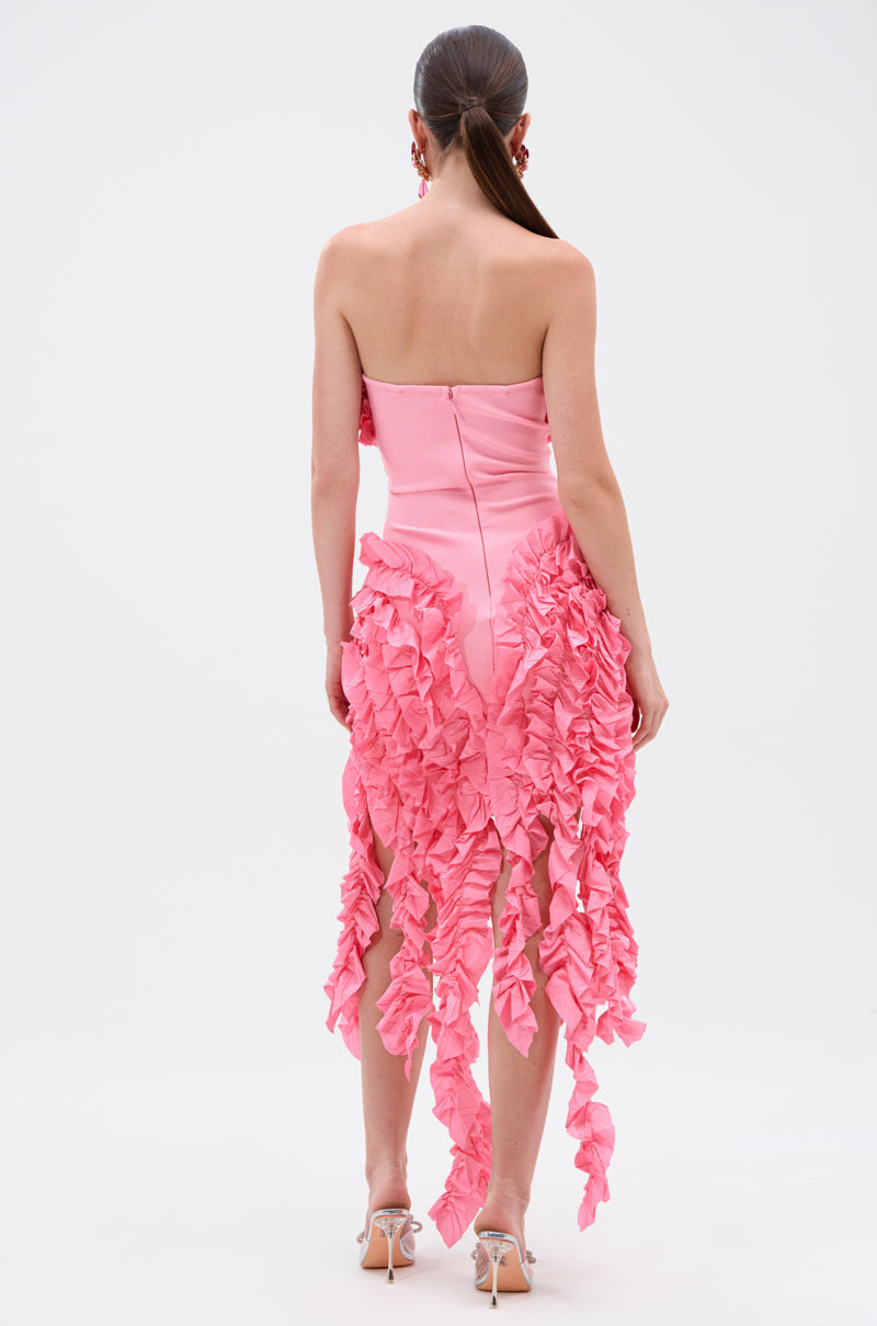 A woman with her hair in a sleek ponytail faces away, wearing the CITY LIGHTS RUFFLE MIDI DRESS IN LIGHT PINK—strapless, fitted, featuring cascading ruffles from waist to knees—and strappy high-heeled sandals.