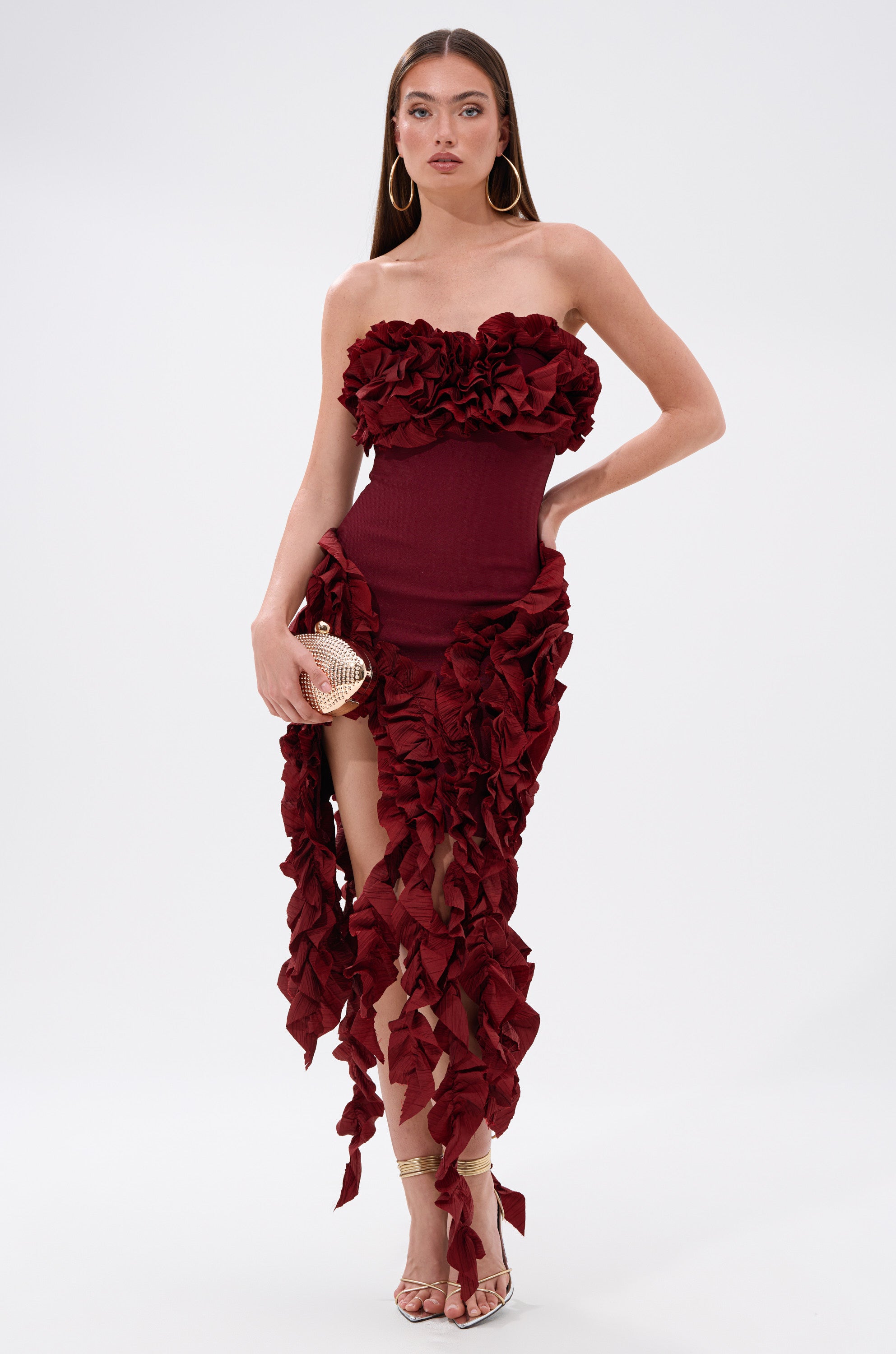 A woman wears the CITY LIGHTS RUFFLE MIDI DRESS IN BURGUNDY, featuring bold ruffle details. She accessorizes with a gold clutch, hoop earrings, and gold heels, standing against a plain white background.