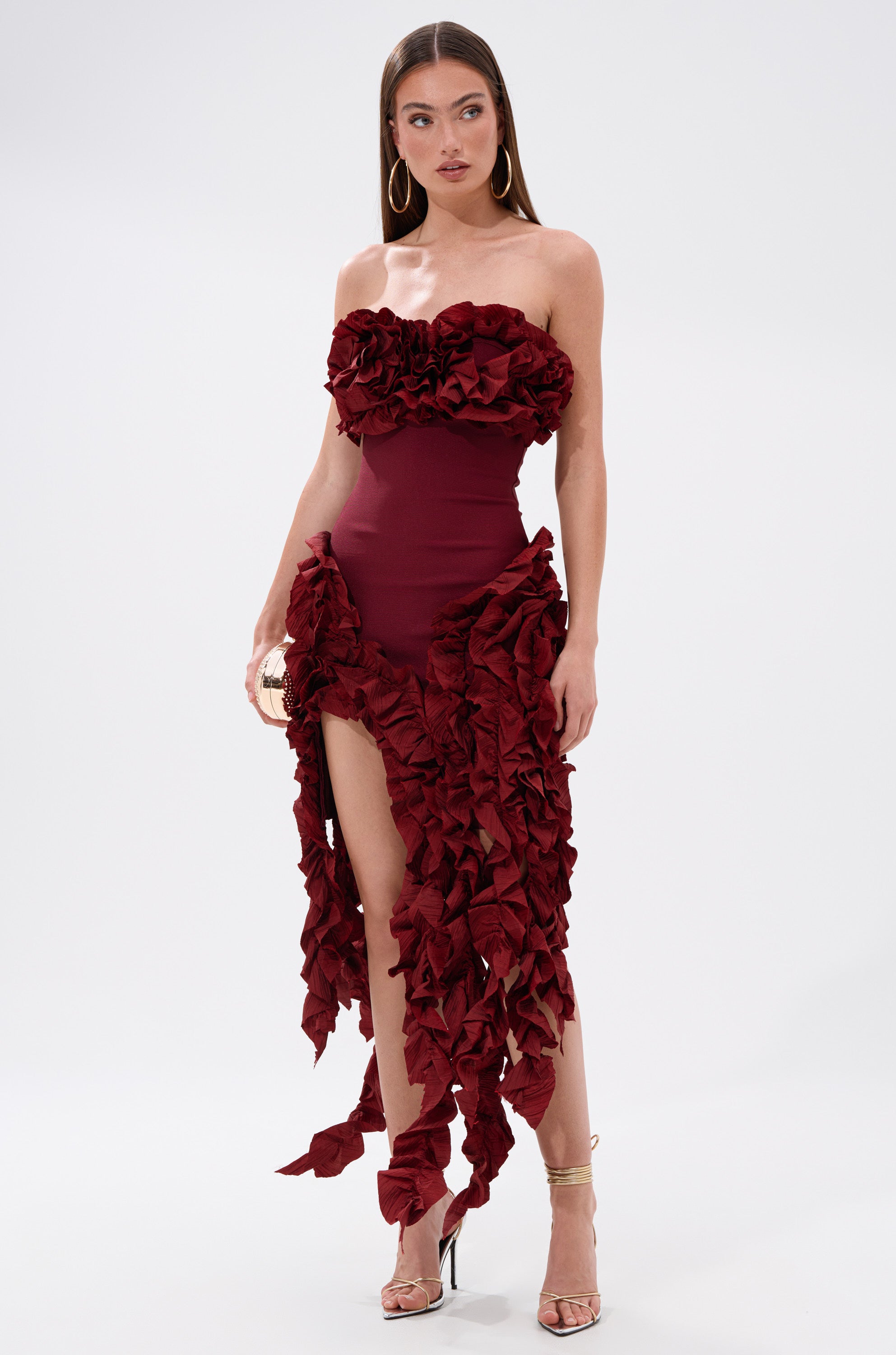 A woman stands against a plain background wearing the CITY LIGHTS RUFFLE MIDI DRESS IN BURGUNDY—a strapless, form-fitting dress with bold textured ruffles—paired with gold hoop earrings and strappy heels.