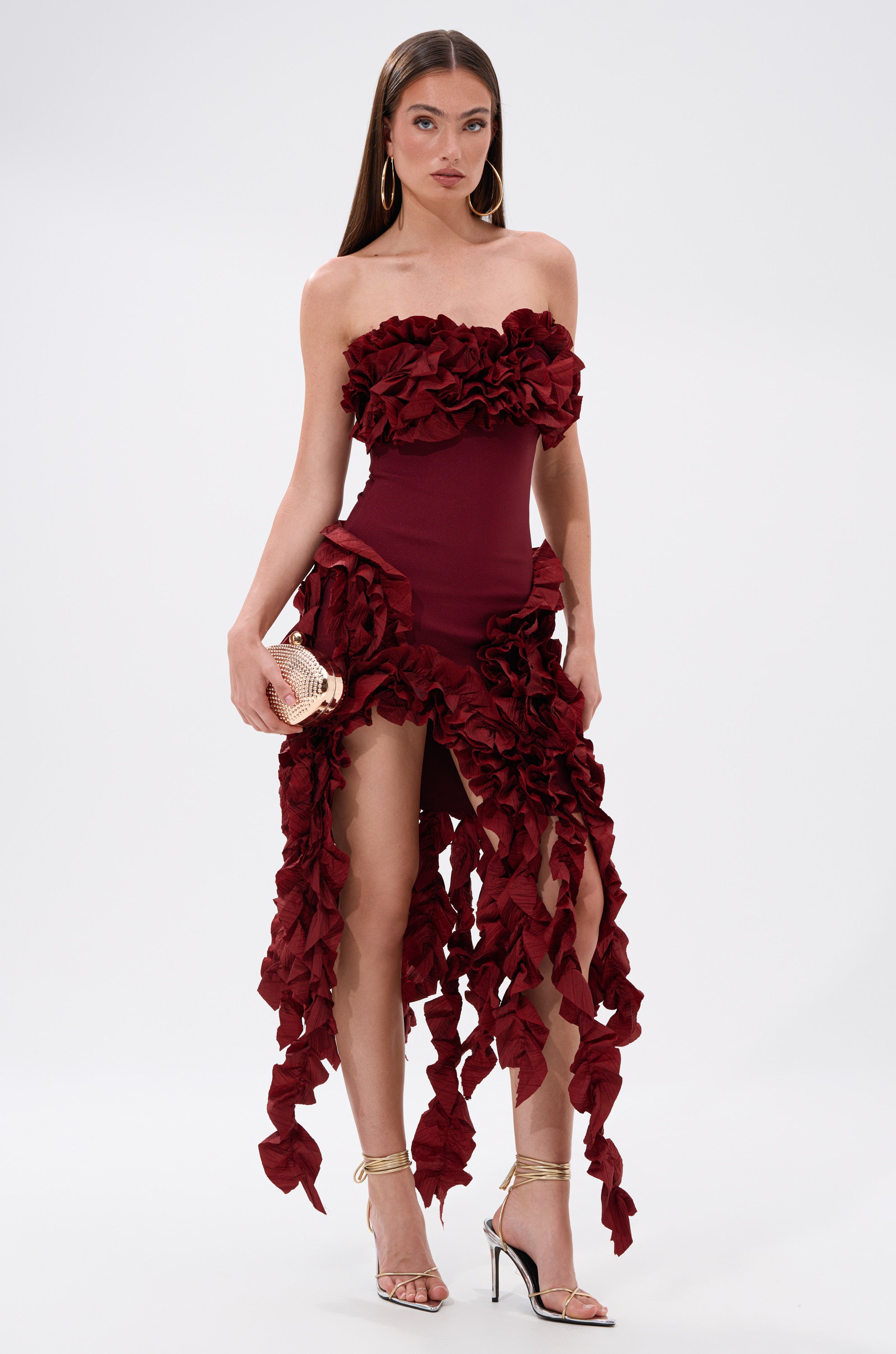 A woman stands against a white background wearing the CITY LIGHTS RUFFLE MIDI DRESS IN BURGUNDY, featuring strapless design and dramatic ruffles. She holds a small clutch and wears gold high-heeled sandals.