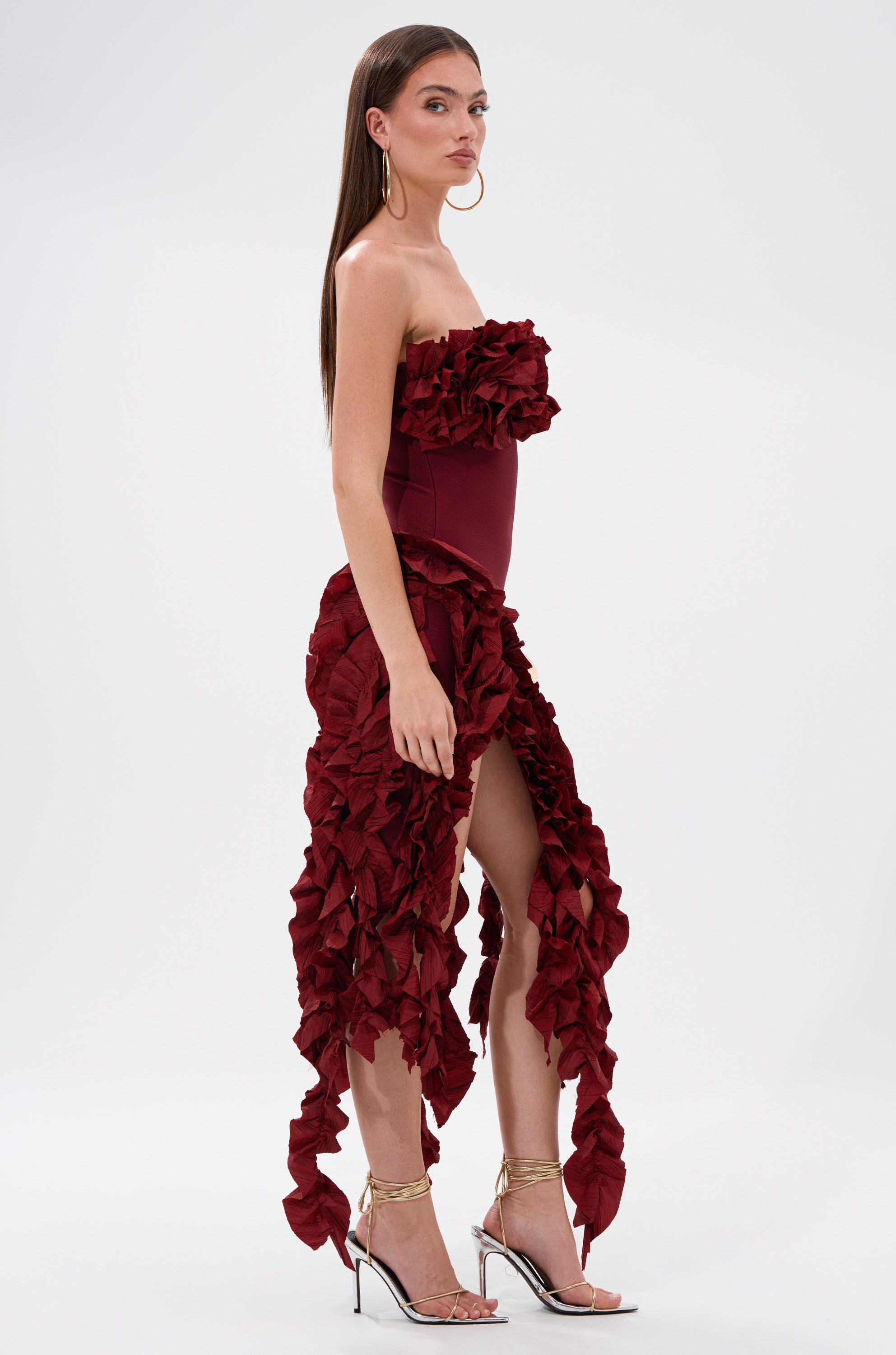 A woman stands in profile wearing the CITY LIGHTS RUFFLE MIDI DRESS IN BURGUNDY, a strapless, ruffled piece with textured layers and a high slit. She pairs it with hoop earrings, silver heels, and has straight long brown hair.