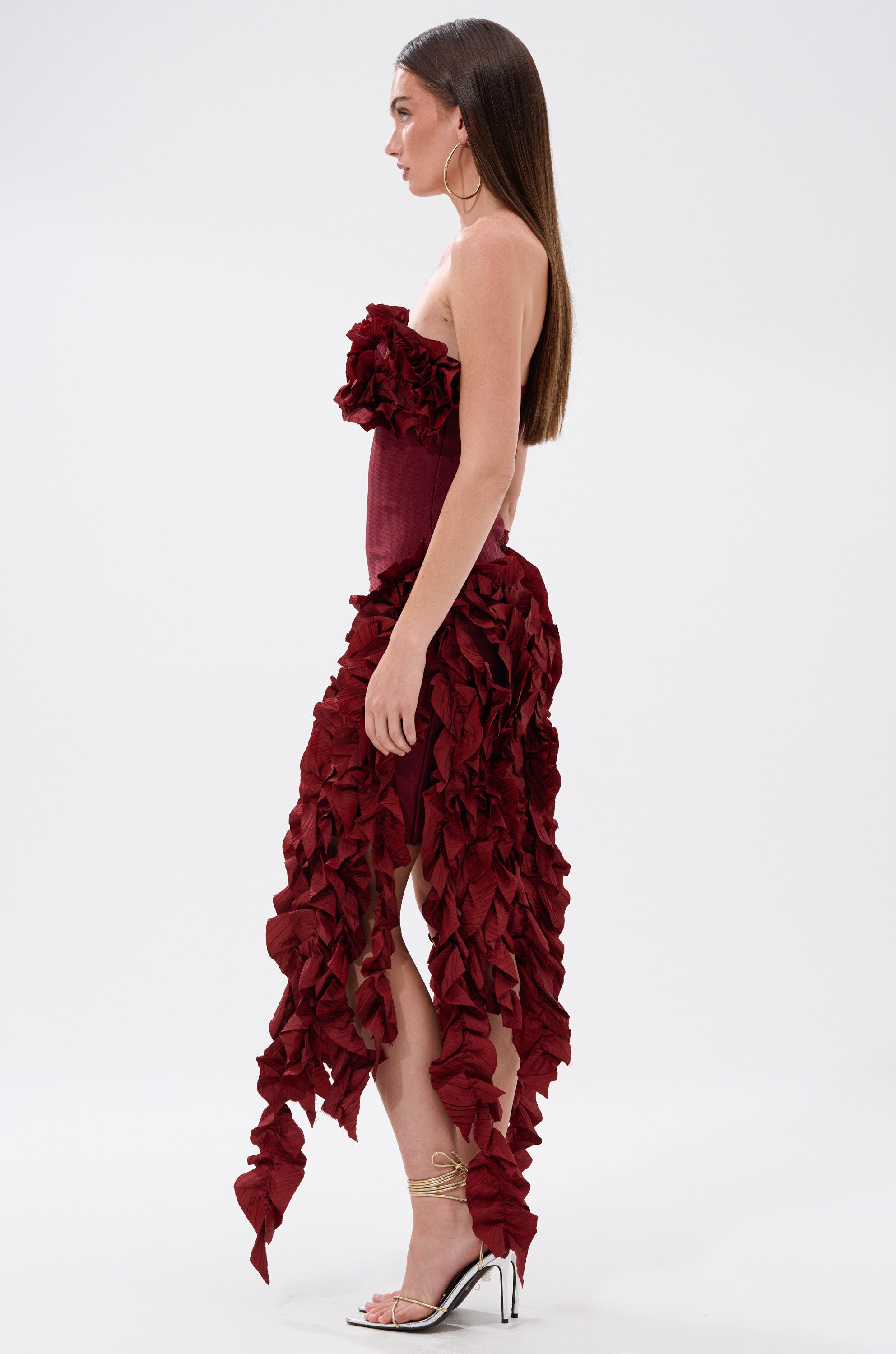 A woman stands in profile wearing the CITY LIGHTS RUFFLE MIDI DRESS IN BURGUNDY, a strapless cocktail dress with cascading ruffled, leaf-like details. She has long brown hair, hoop earrings, and strappy high heels.