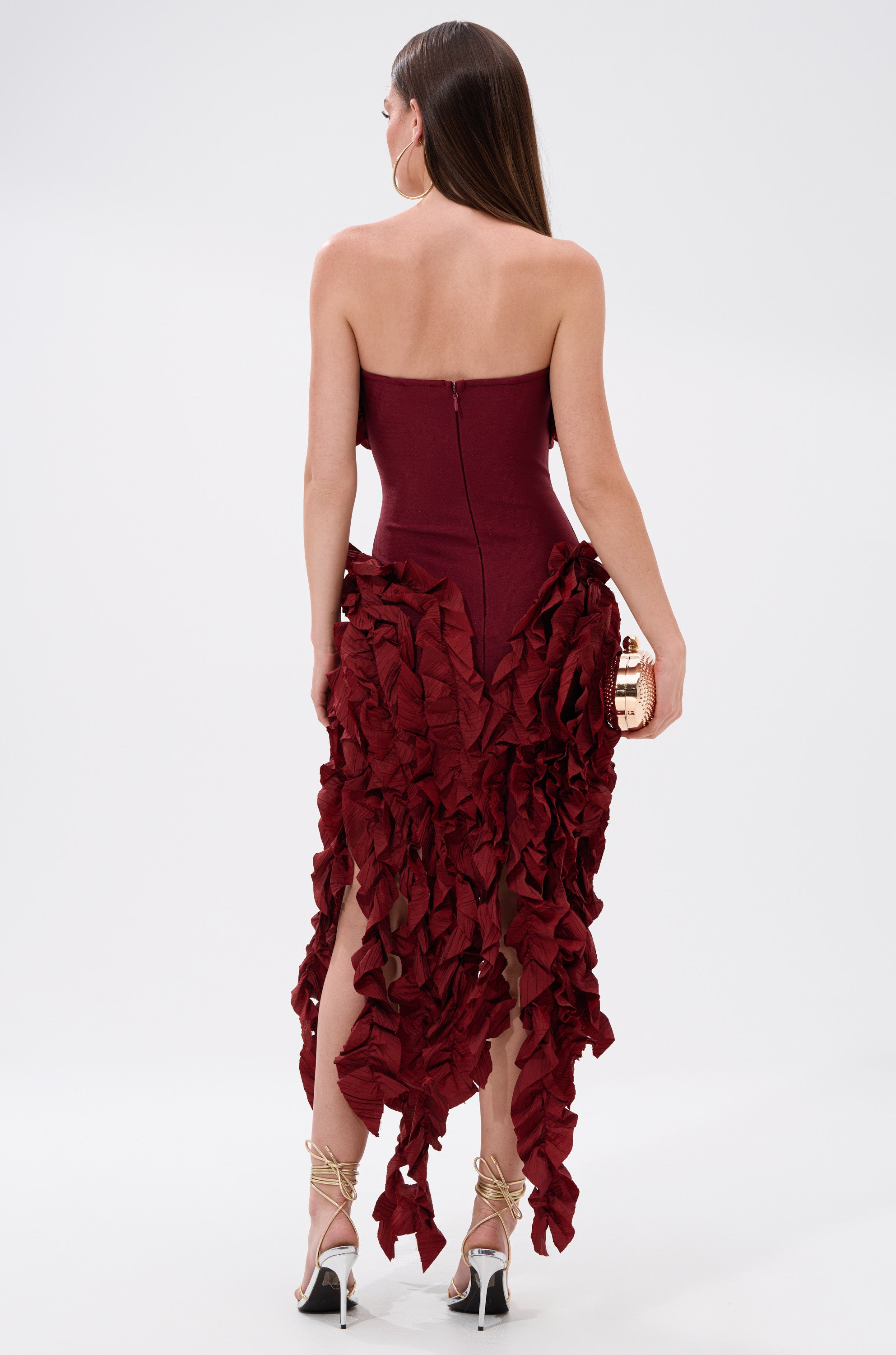 A woman stands facing away in the CITY LIGHTS RUFFLE MIDI DRESS IN BURGUNDY, a strapless bandage dress with an asymmetrical ruffle midi skirt. She holds a gold clutch and wears strappy heels, her long straight hair cascading down her back.