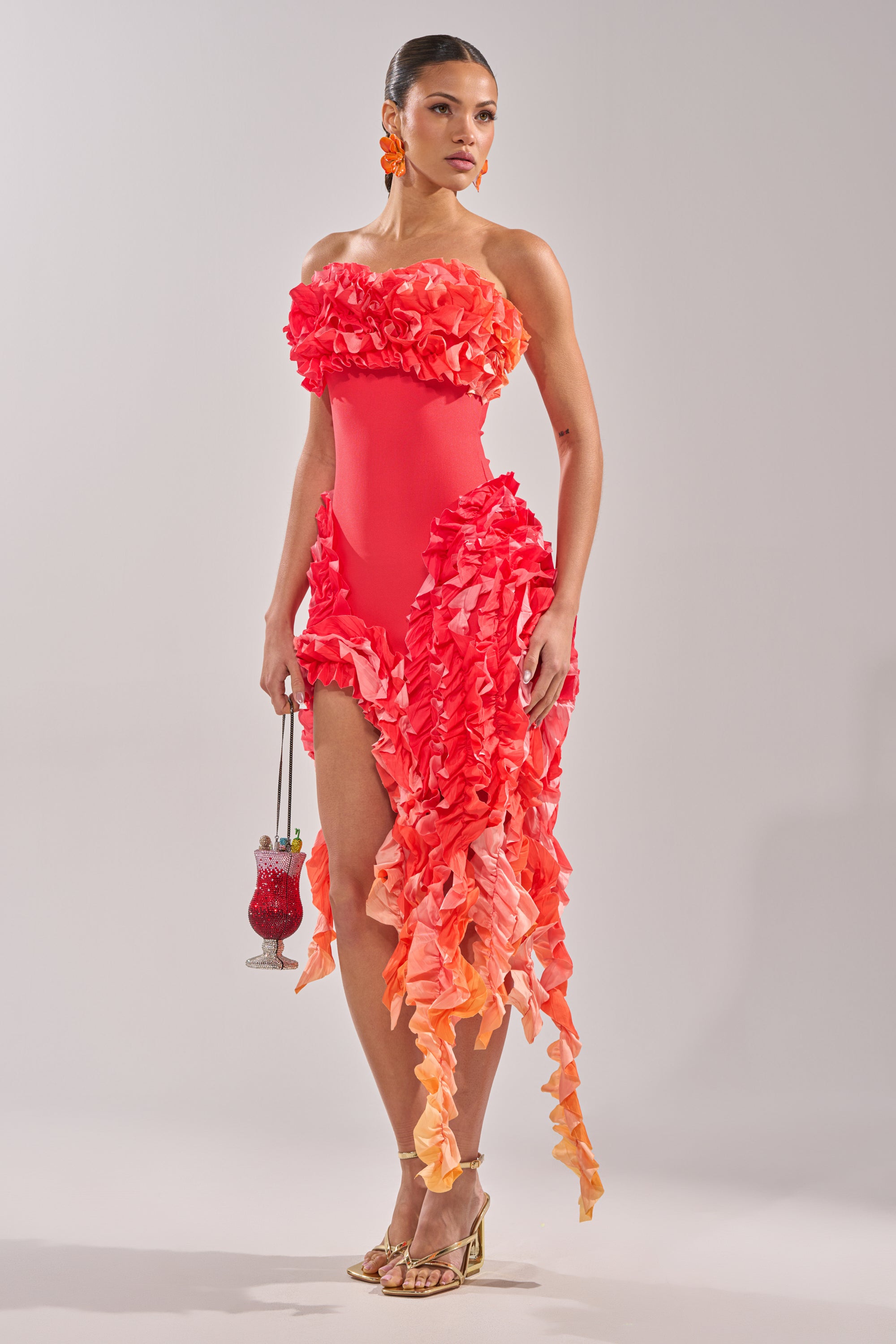 A woman wearing the CITY LIGHTS RUFFLE MAXI DRESS IN RED OMBRE, gold heels, and orange earrings holds a pink beaded cocktail glass-shaped handbag while standing against a plain background.